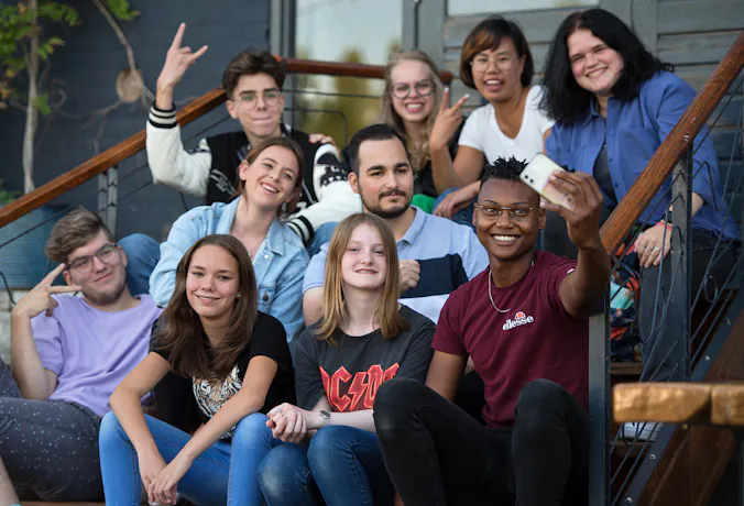 Group of cheerful young people sitting on a staircase, with one person in the front taking a selfie.