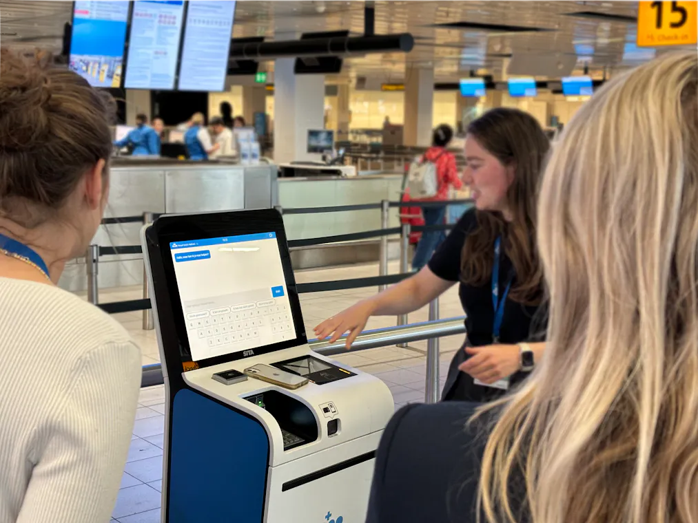 KLM-medewerker die bij een kiosk op Schiphol twee mensen uitleg geeft.