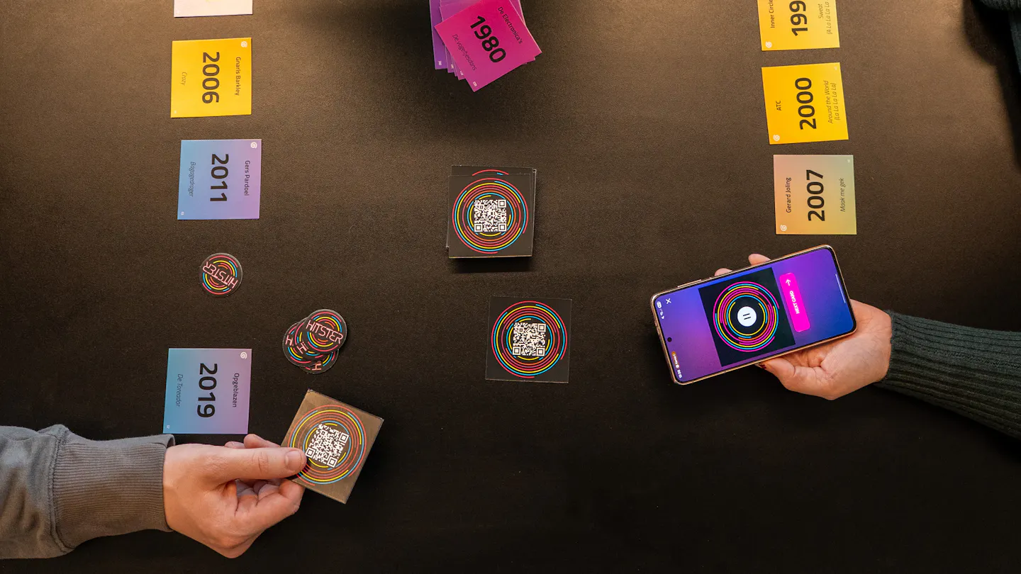 Hitster gezelschapsspel op een tafel met de handen van twee spelers, waarbij de ene een speelkaartje vasthoudt en de ander een telefoon met de app.