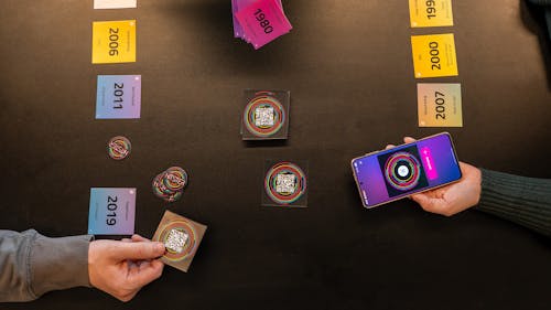 Hitster bordspel op een tafel met de handen van twee spelers, waarbij de ene een speelkaartje vasthoudt en de ander een telefoon met de app.