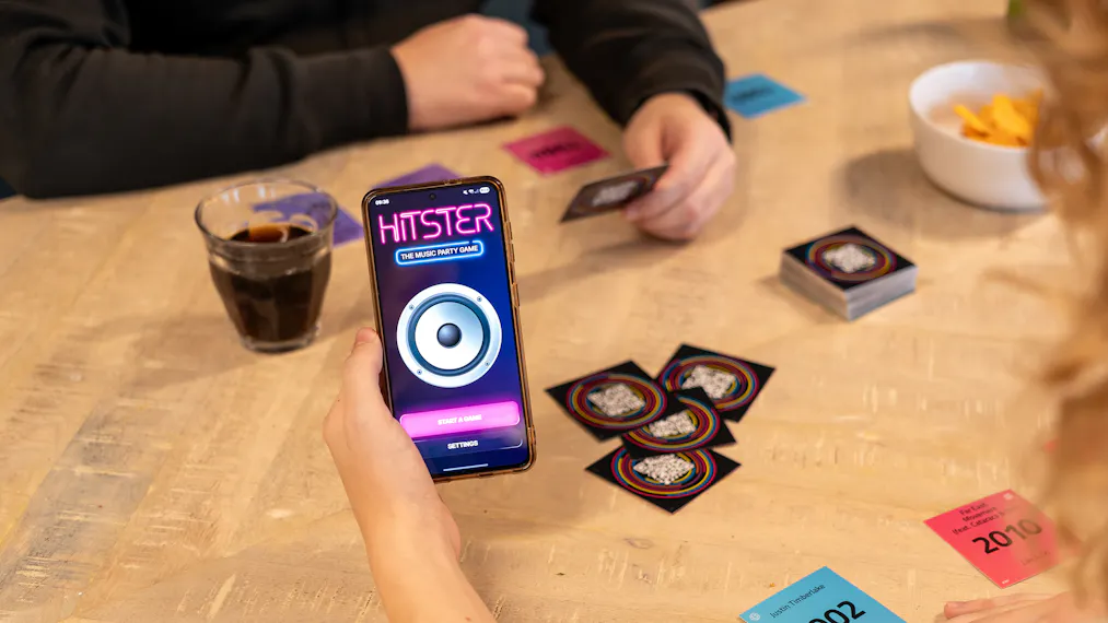 Hitster gezelschapsspel op een tafel met de handen van twee spelers, waarbij de ene een speelkaartje vasthoudt en de ander een telefoon met de app.