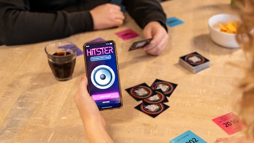 Hitster bordspel op een tafel met de handen van twee spelers, waarbij de ene een speelkaartje vasthoudt en de ander een telefoon met de app.