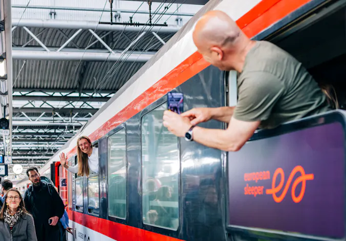 Train from outside with a man leaning out of a window to take a picture of a woman waving from another window.
