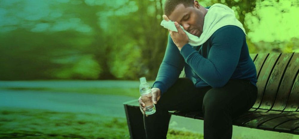 Man sitting on a park bench, drinking water and wiping sweat from his face with a towel after a run.
