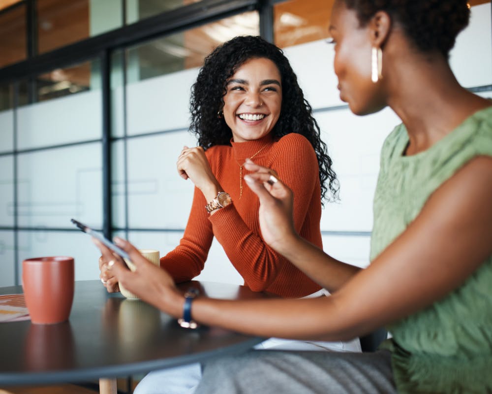 two women sitting together talking and laughing with a phone in hand and a coffee cup on the table in front of them