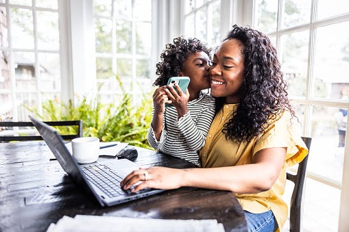 Woman working on a laptop at a table while a young child kisses her on the cheek in a bright home setting.