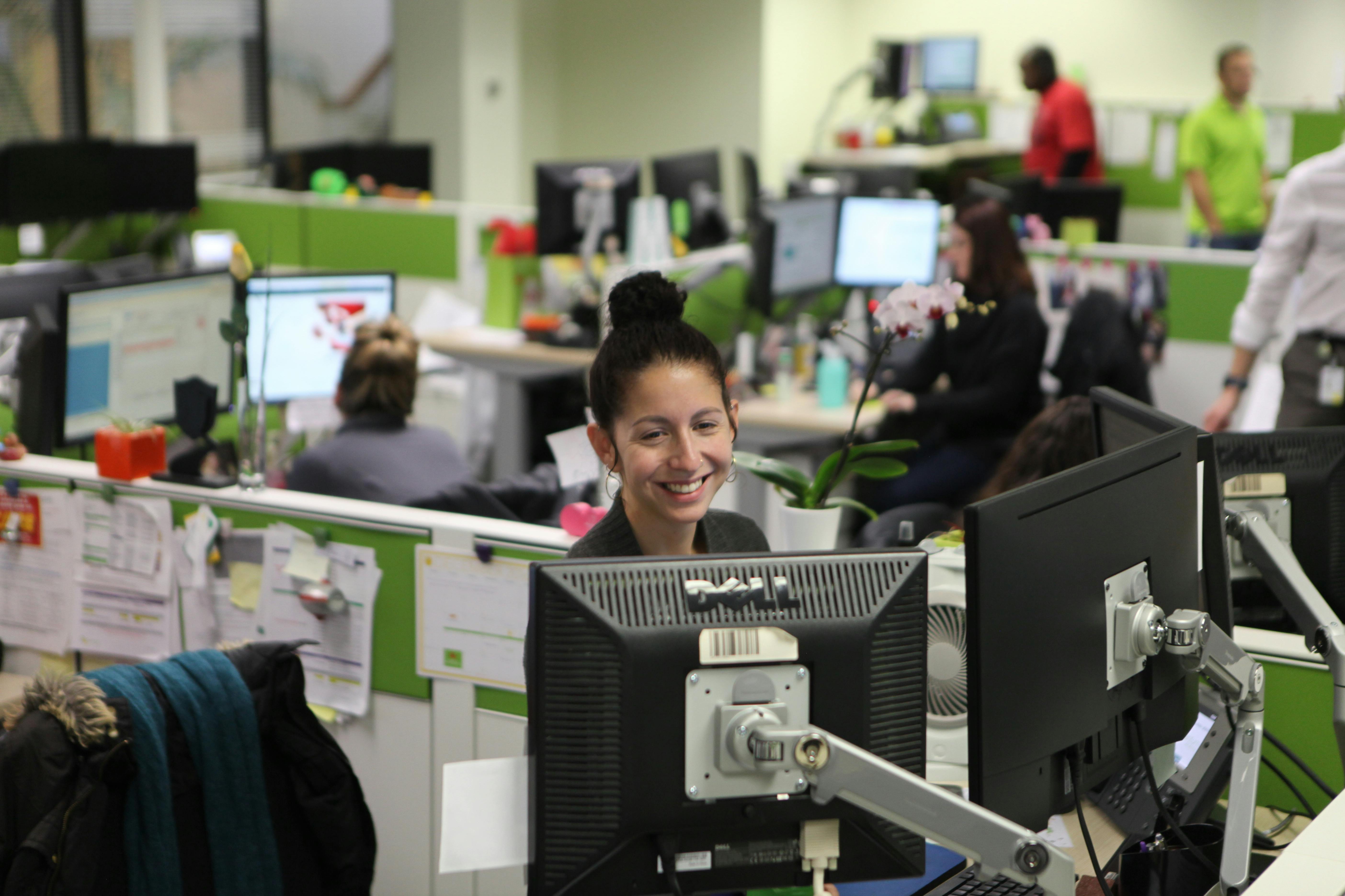 Employees working at computer stations in an open-plan office, with multiple desks, monitors, and people collaborating in the background.