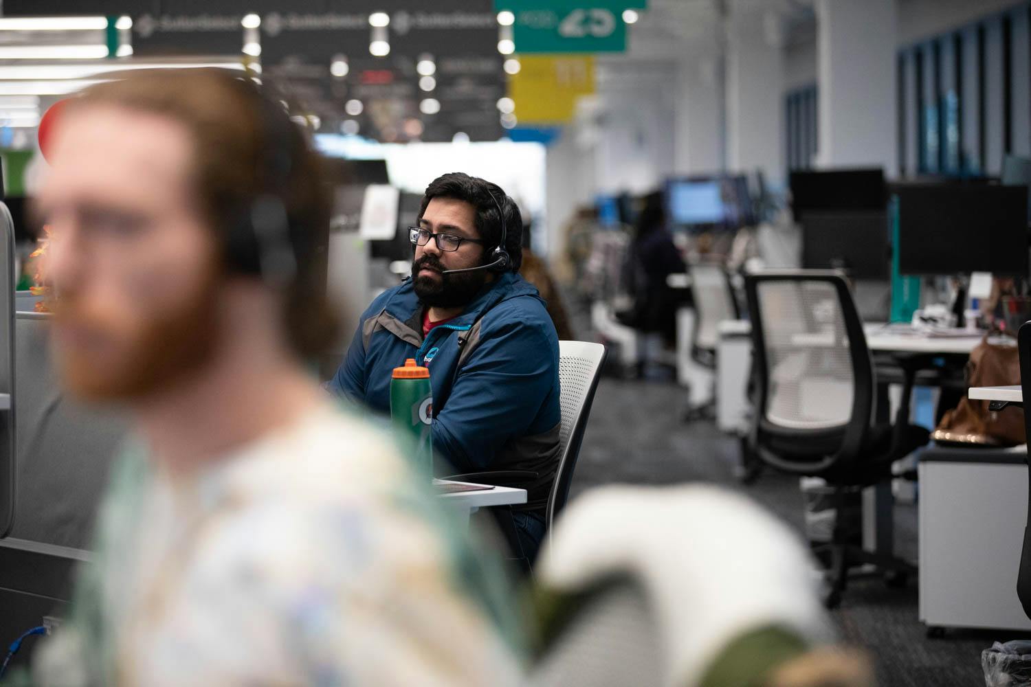 People working at desks in a modern open-plan office, with one person in focus wearing headphones and others moving or working in the background.