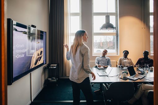 A woman presenting to a small group in a conference room, standing beside a large wall-mounted screen while colleagues sit at a table listening.