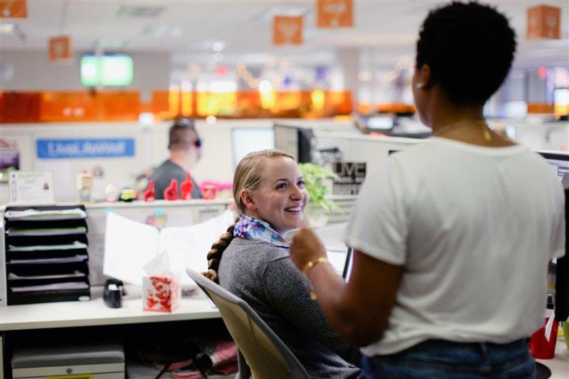 Two coworkers talking in an open-plan office. One person sits at a desk and smiles while turning toward a colleague who is standing nearby, with desks, computers, and office signage visible in the background.