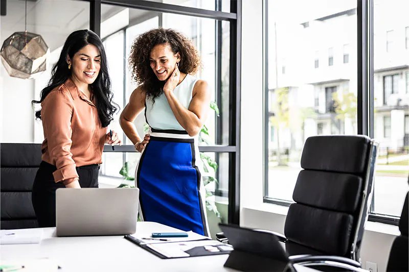 Two professionals standing in a modern office near a desk with a laptop, collaborating during a meeting.