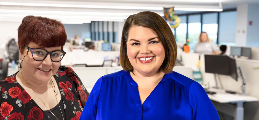 Two women smiling in a modern open-plan office. One wears glasses and a black top with red floral patterns, the other wears a bright blue blouse; desks, computers, and coworkers are visible in the softly blurred background.