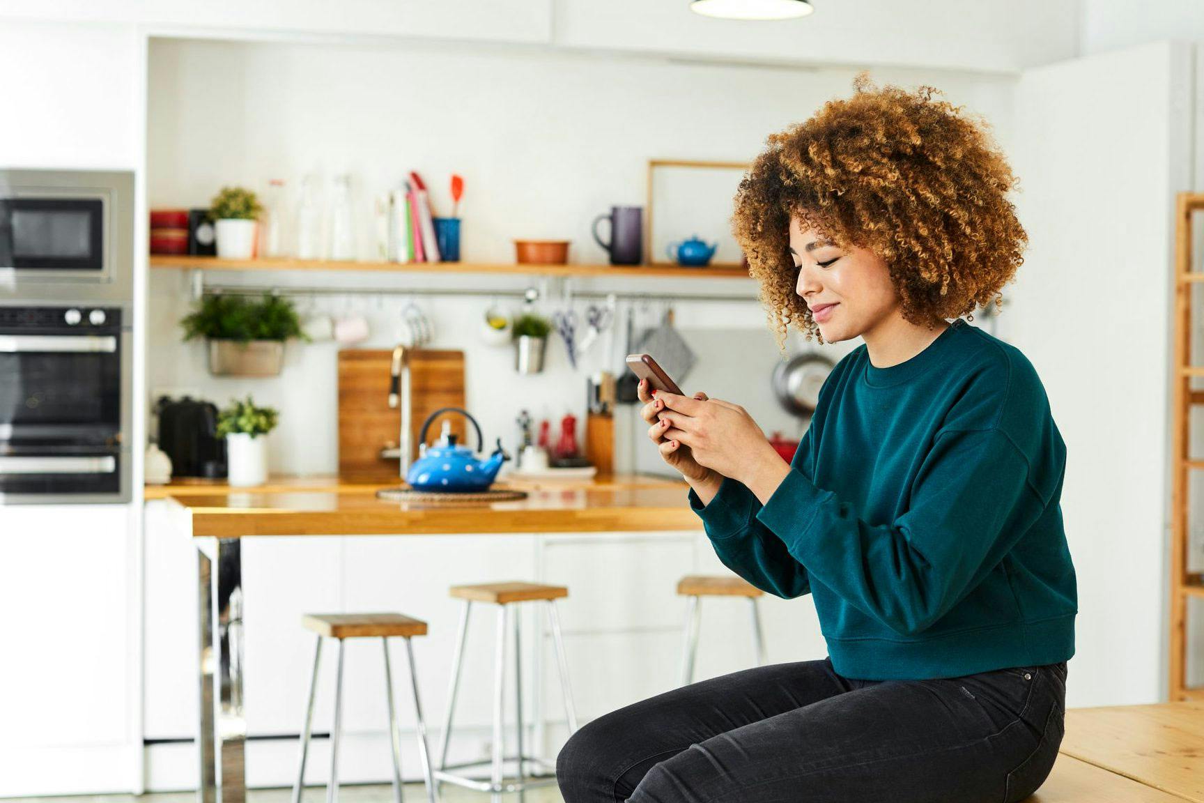 A woman sitting on a wooden bench in a modern kitchen, looking at her phone.