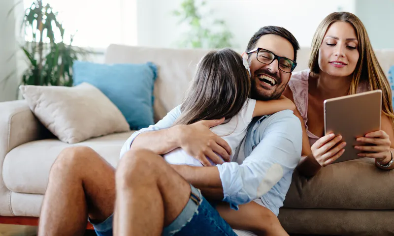Man sitting on ground in front of couch hugging young girl in his lap while woman lays behind them on couch smiling at a tablet.