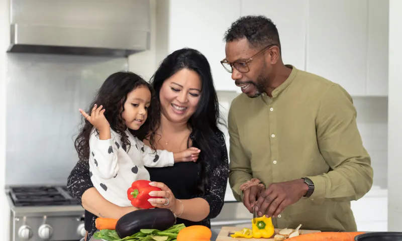 Father standing next to a mother holding a child while he chops vegetables in the kitchen