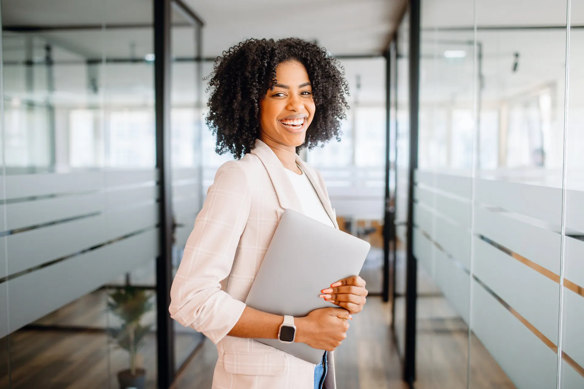 A woman in a light-colored suit holding a laptop in an office hallway.