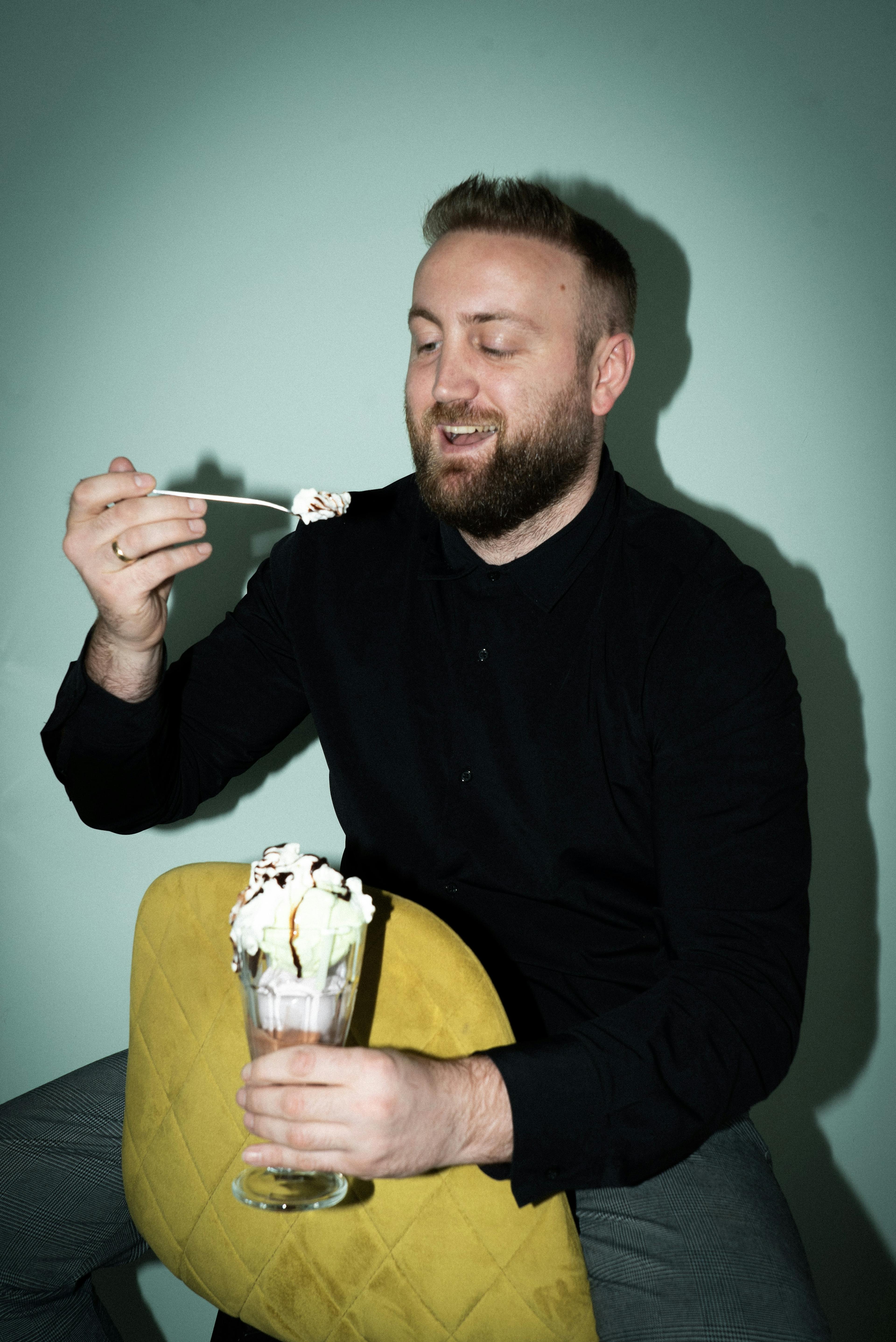 A person is seated, holding a spoonful of ice cream from a sundae glass filled with ice cream. They are wearing a black long-sleeved shirt and grey pants, seated on a yellow cushioned chair against a light teal wall.