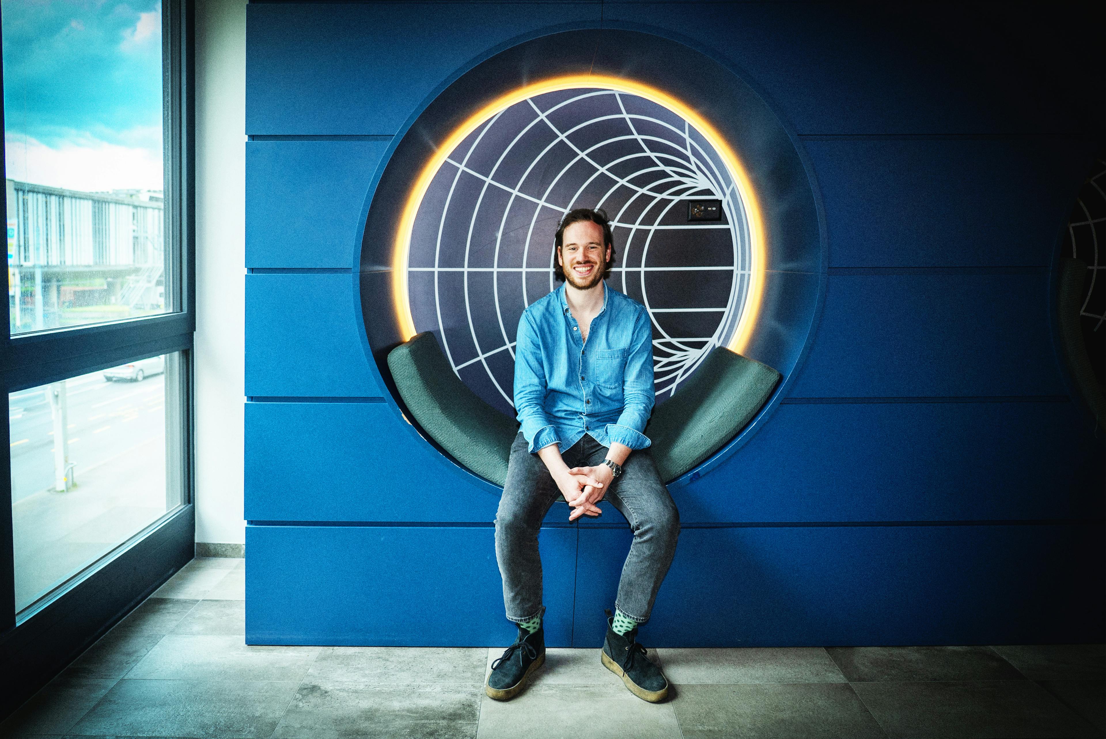 A person is seated in a modern, circular blue chair with white lighting accents, against a vibrant blue wall adorned with a unique lighting design.