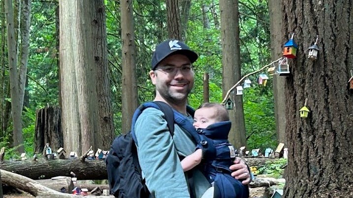 Headshot of Ryan Fry. He is wearing a blue button up shirt, purple plants, a baseball cap, a backpack, and is holding his infant child