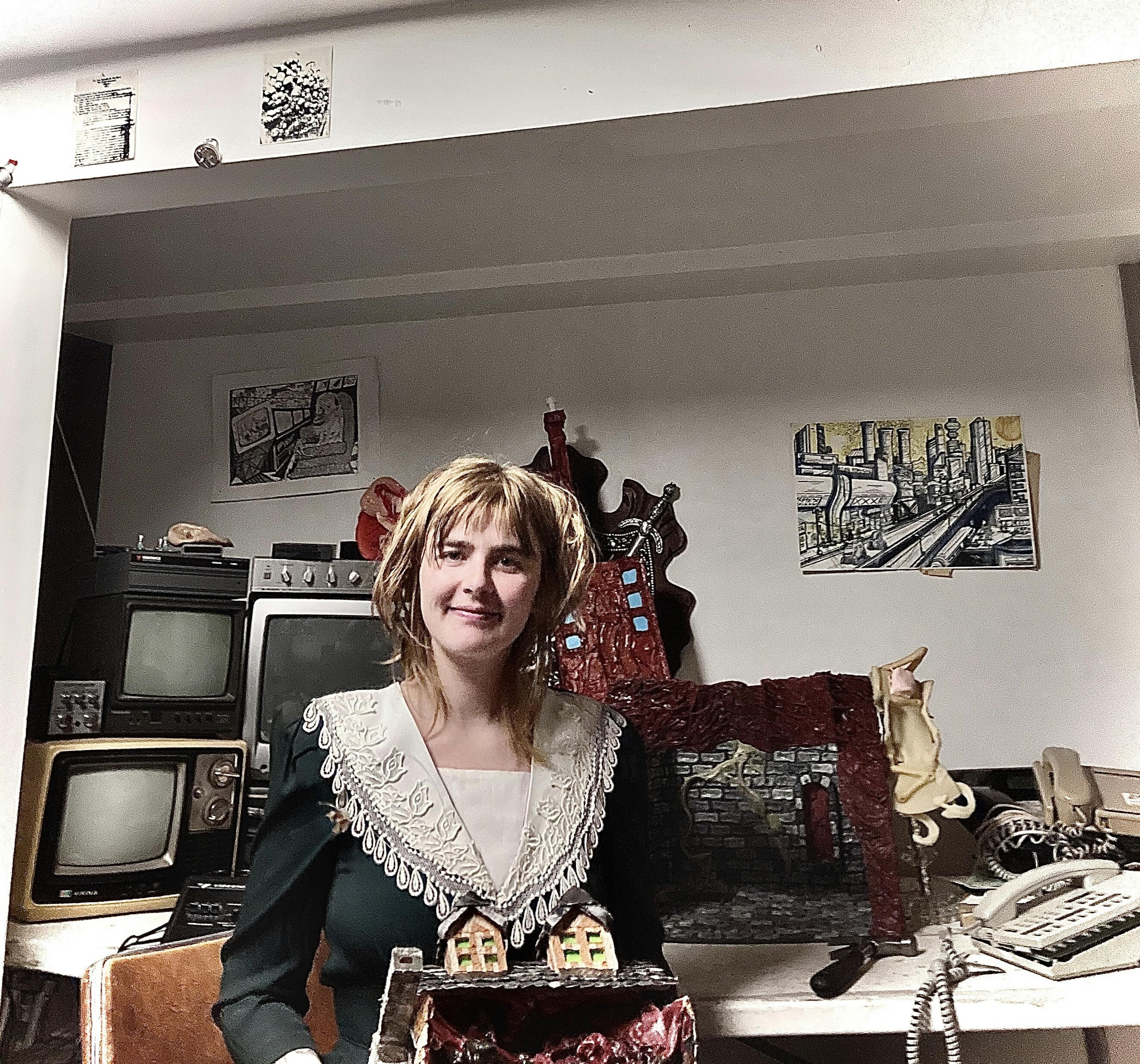 A photograph of Kayla Jane Macneill. She is looking at the camera, holding a small diorama of a house, whilst behind her is a desk cluttered with old crt tvs and telephones. 

She has shoulder length blonde hair, brown eyes, and is wearing a lacy-collared sailor suit. 