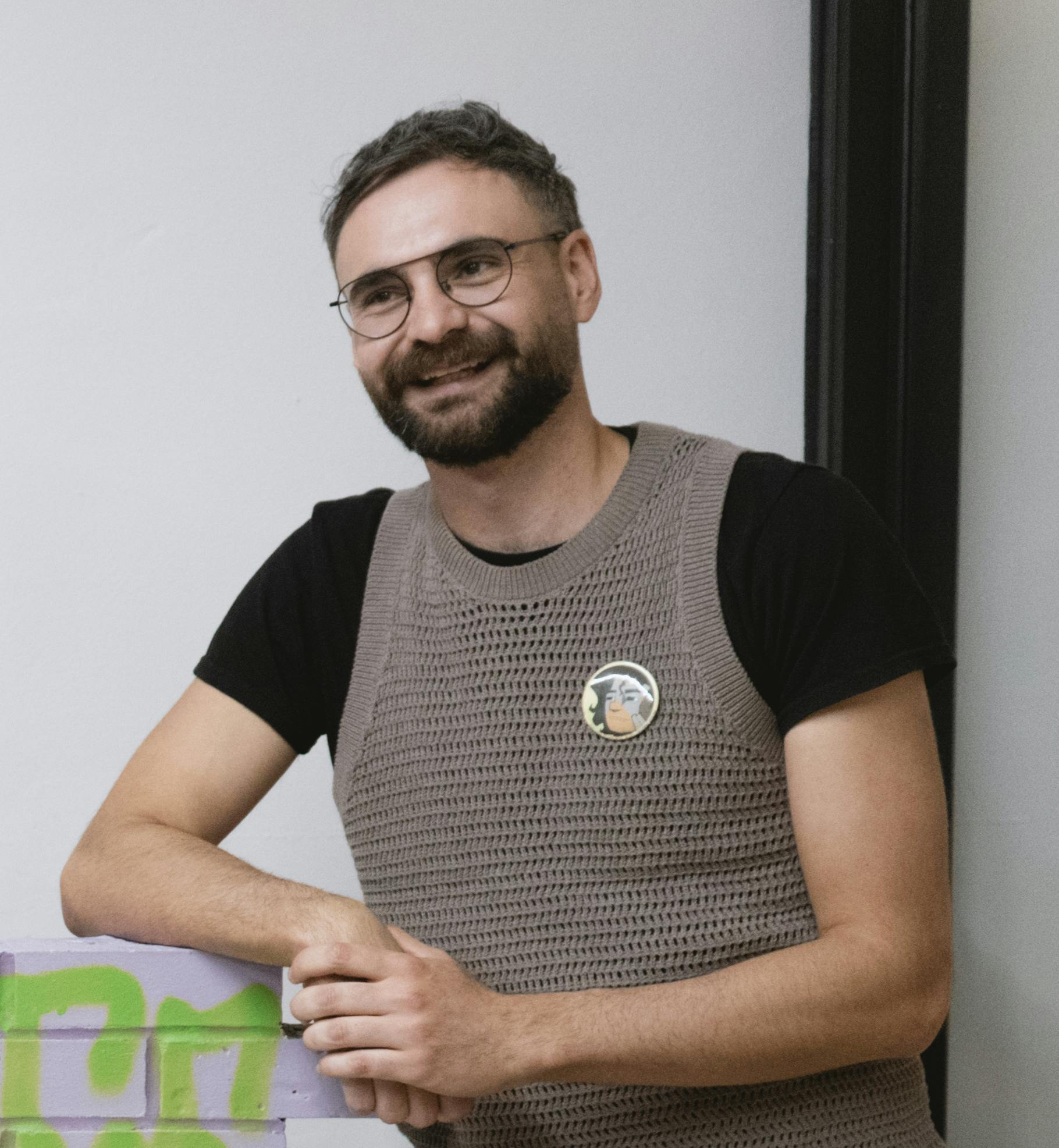 Ryan Von Hagen smiling and looking just off camera. Ryan has short brown hair, a beard and moustache (close-cut), wears round-rimmed glasses, and is wearing a black t shirt with a taupe knit vest overtop. He is leaning on a wall. 