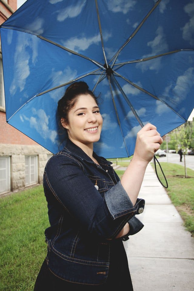 Natasha Jensen headshot. She is looking at the camera while holding an umbrella that has clouds on it. She has brown hair tied up in a bun, and is wearing a denim jacket