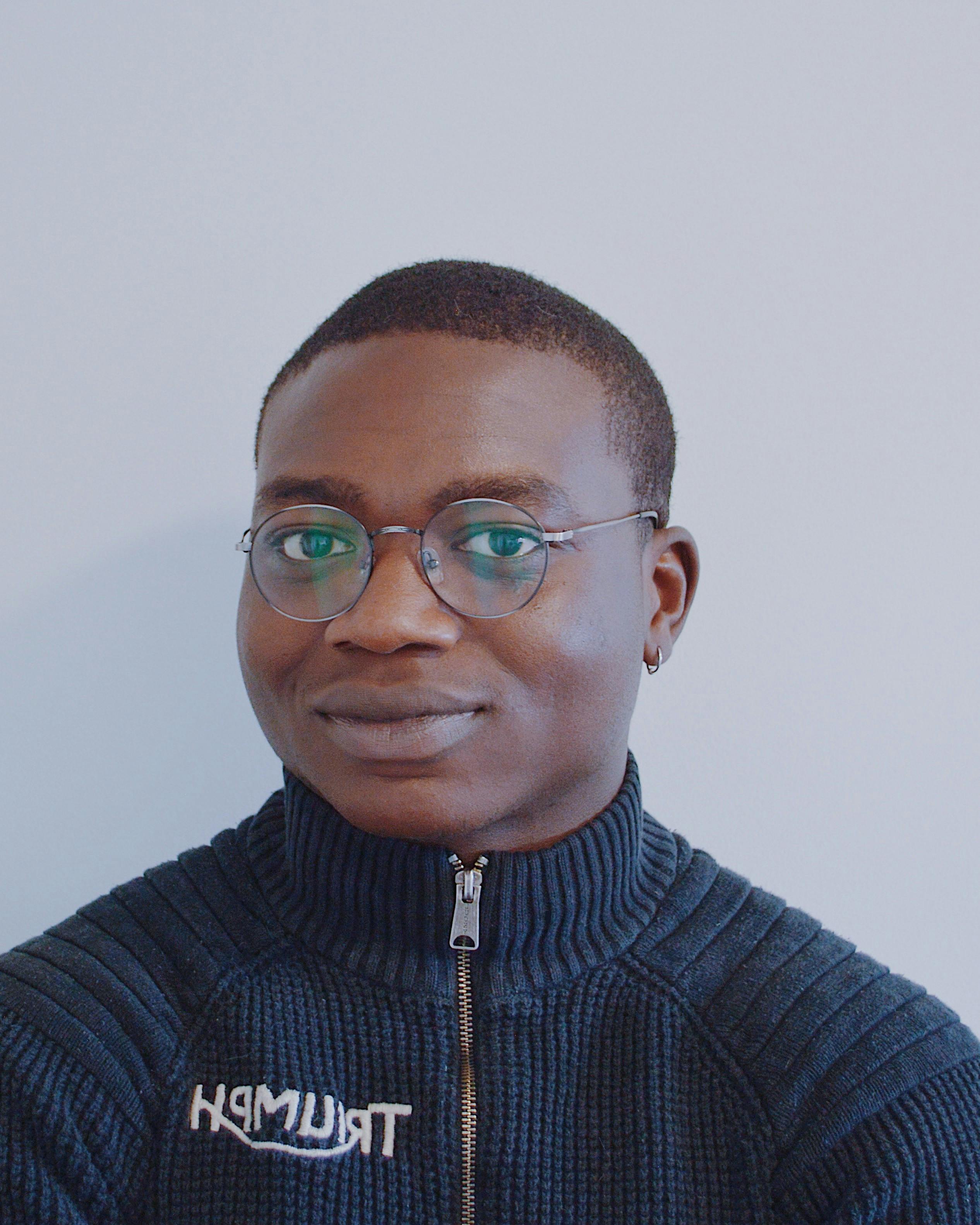 headshot of Mide Kadiri. Mide has black skin, short black hair, and dark brown eyes. He has an earring in his left ear and is wearing glasses. He's wearing a navy-blue turtleneck with a zipper in the front