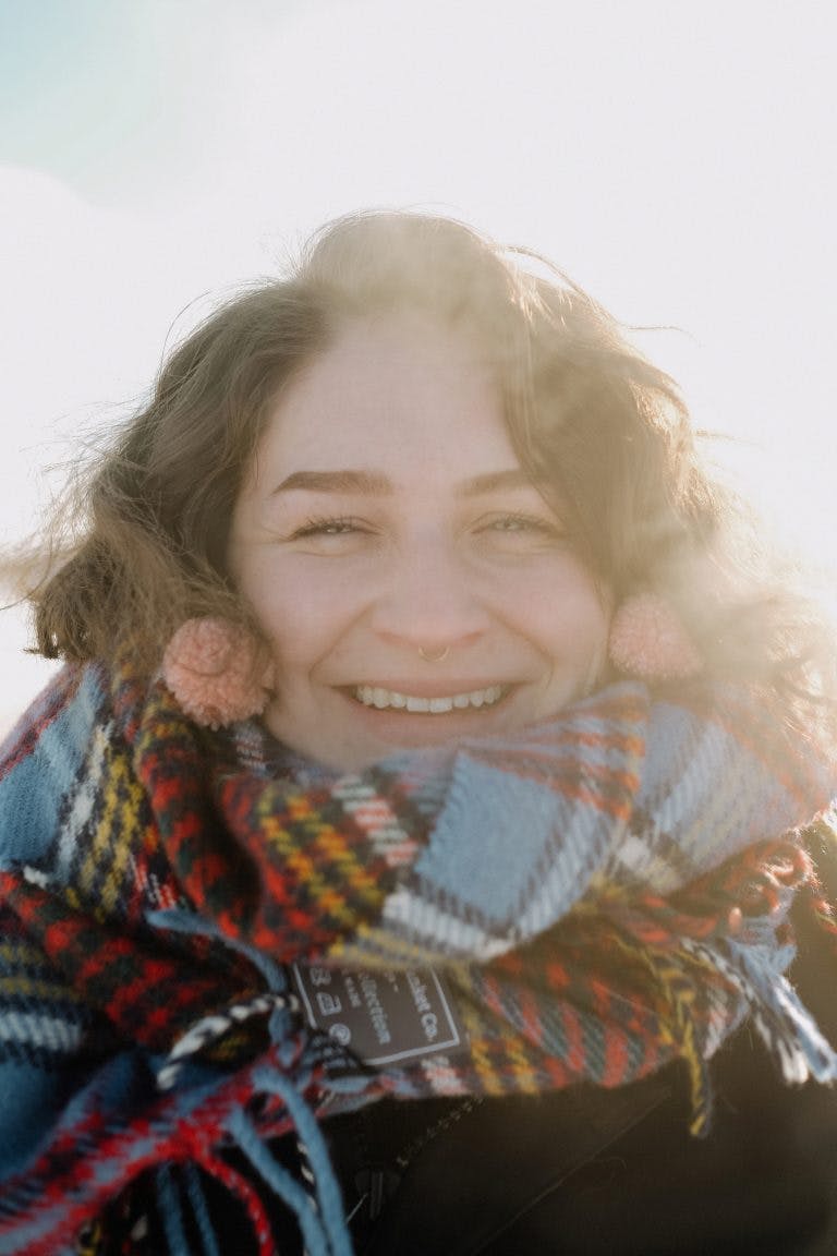 Abbey Bennet is smiling towards the camera. She has brown wavy hair, and is wearing a plaid scarf and pompom earrings. 