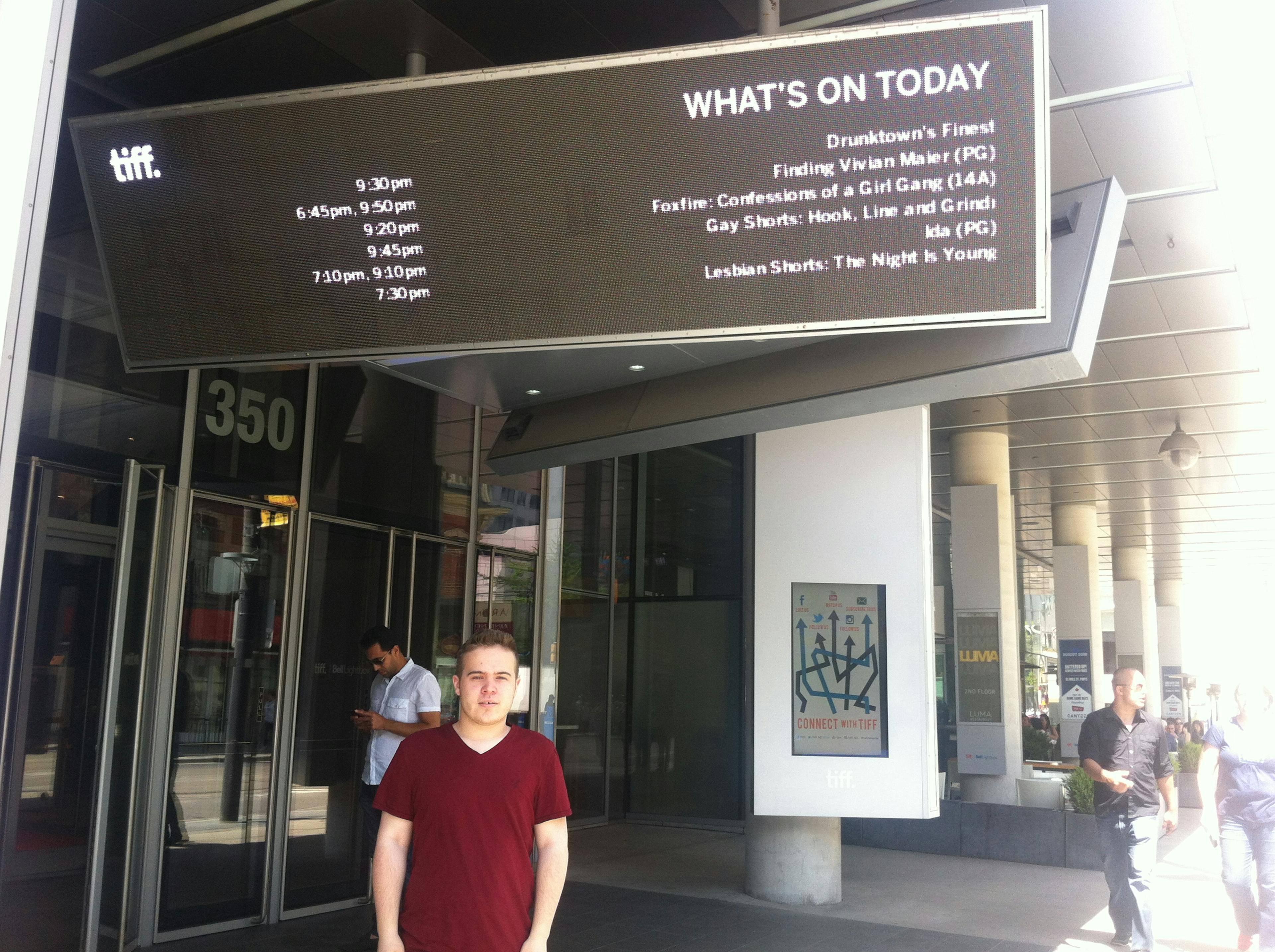 Image of a younger man, wearing a red t shirt, underneath a tiff sign that reads "What's on today" with various screenings