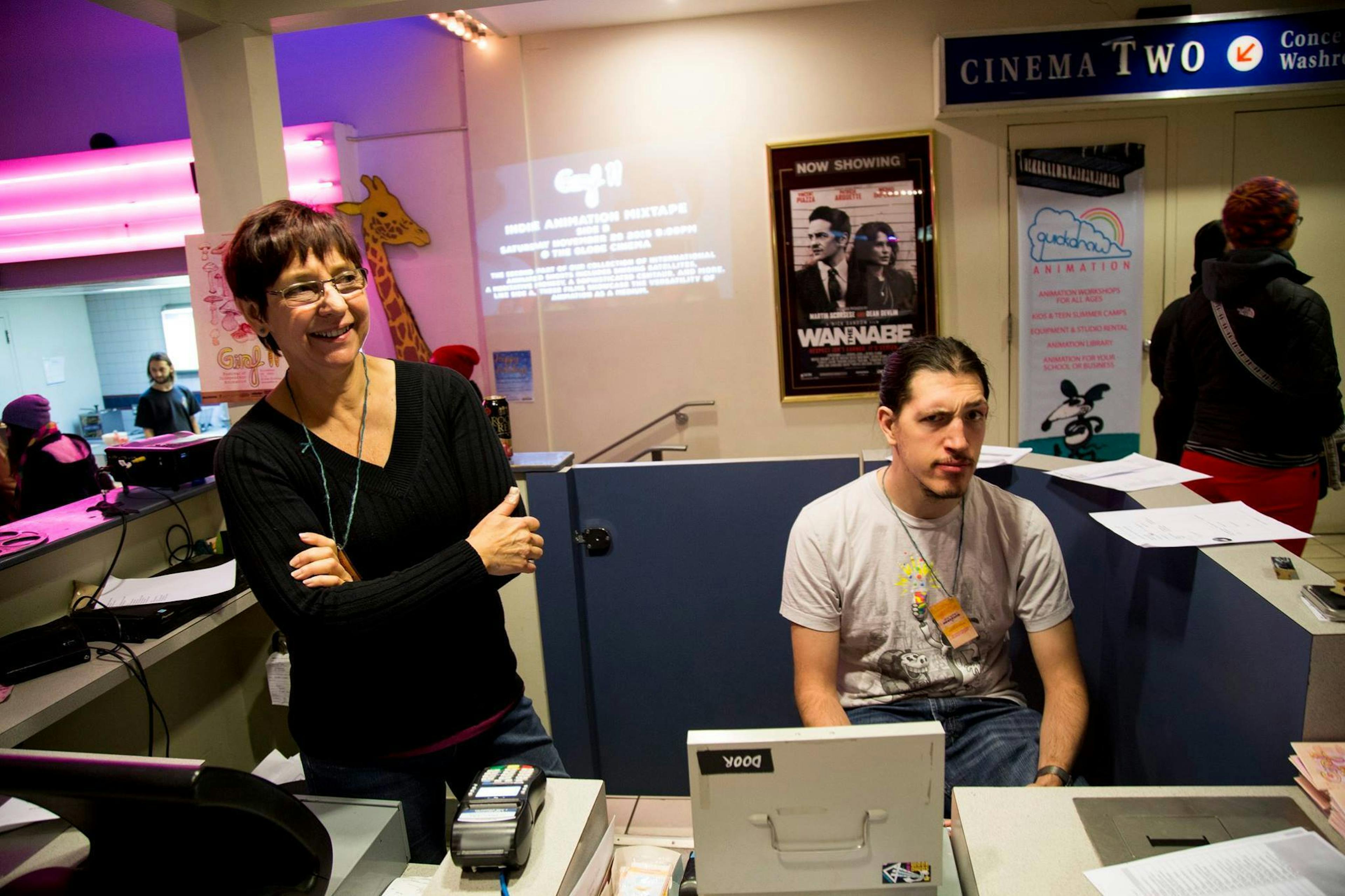 An image of a front desk at a movie theatre. There are two people behind the desk, one of which is Will Walton. Will has long hair tied back into a pony tail, has a short mustache and beard, and is comically raising just one eyebrow. He is sitting, wearing a grey tshirt and a GIRAF11 pass around his neck. 