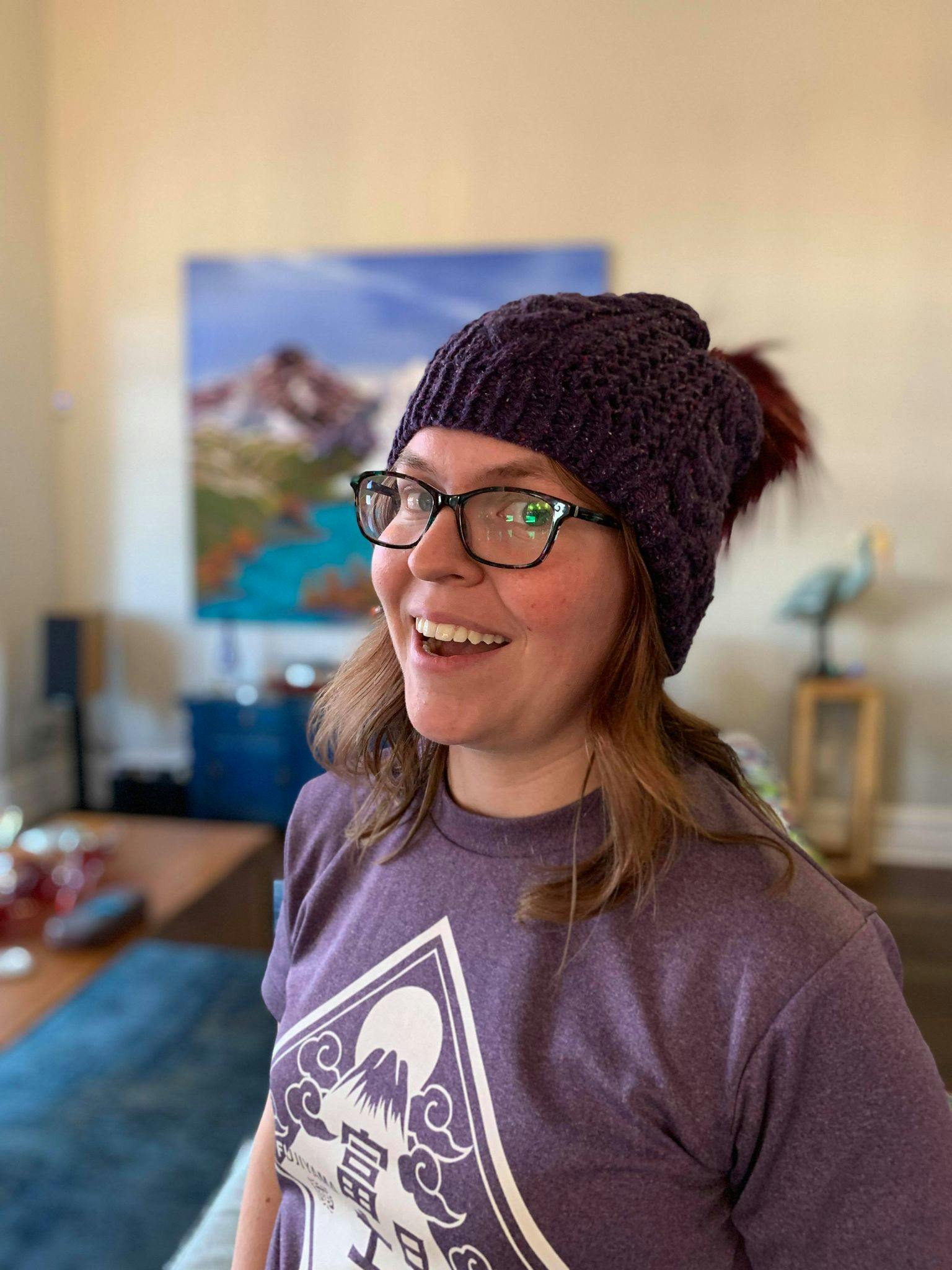 A picture of Theresa Pinhey smiling at the camera. Theresa has long brown hair, green eyes, and she's wearing dark blue glasses. She is wearing a purple knit hat and a purple t-shirt