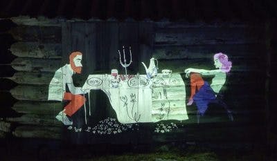 A projected image onto a  cabin wall, two people sitting at a table that has a glass of wine, a candelabra, a tea pot, and assorted plates. There is a tablecloth on the table, but the man on the left looks like he's pulling the table away from the women across from him. He has red hair, a beard, a white shirt and red pants. She has purple curly hair, a white blouse, a blue skirt and red tights
