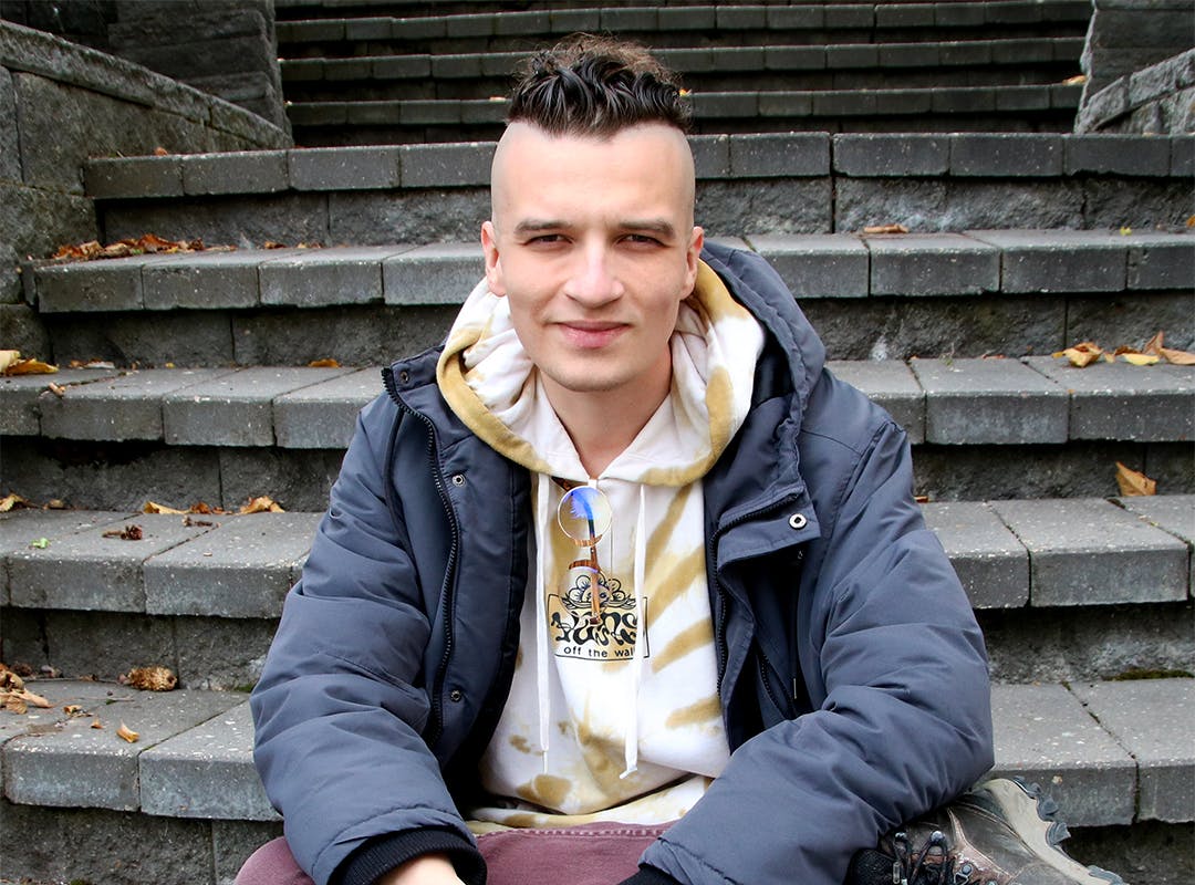 Portrait of Hartley Fiala. He is sitting on some stairs and looking into the camera with a rested smile. He has the side of his head shaved but the top of his hair is dark brown and short. He is wearin ga white and brown hoodie, a winter jacket, burgundy jeans, and hiking boots. His legs are crossed and his hands are clasped, resting on his knee. 