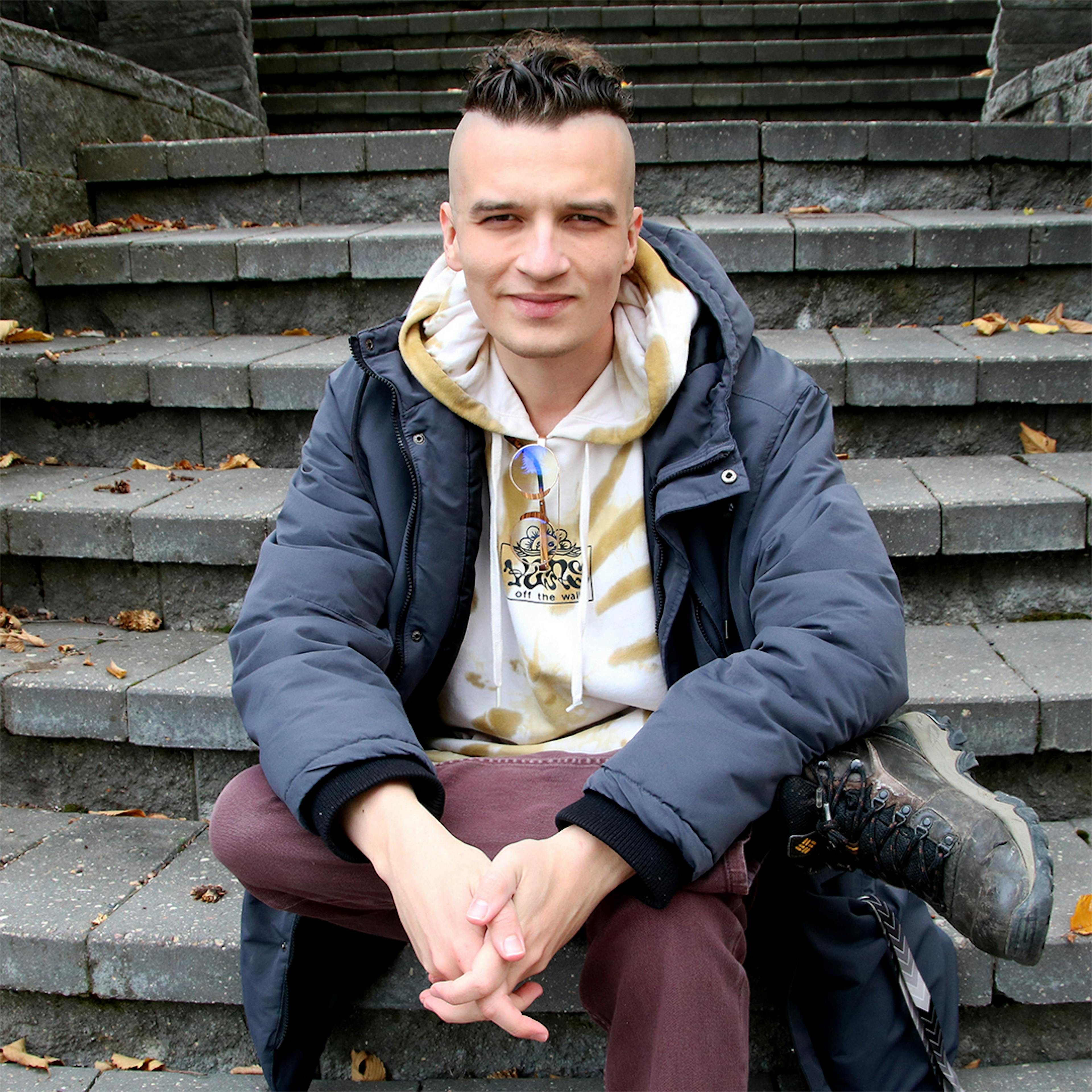 Portrait of Hartley Fiala. He is sitting on some stairs and looking into the camera with a rested smile. He has the side of his head shaved but the top of his hair is dark brown and short. He is wearin ga white and brown hoodie, a winter jacket, burgundy jeans, and hiking boots. His legs are crossed and his hands are clasped, resting on his knee. 