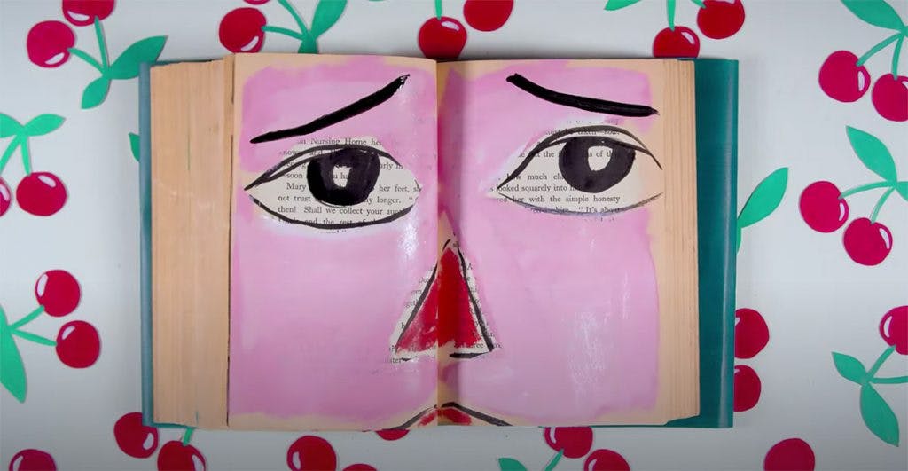 A book sits on a table with a cherry-patterned table cloth. The book lays open, and inside is a painted face covering the words, one eye on each side of the open book with the nose over the spine. The face looks lonely. 