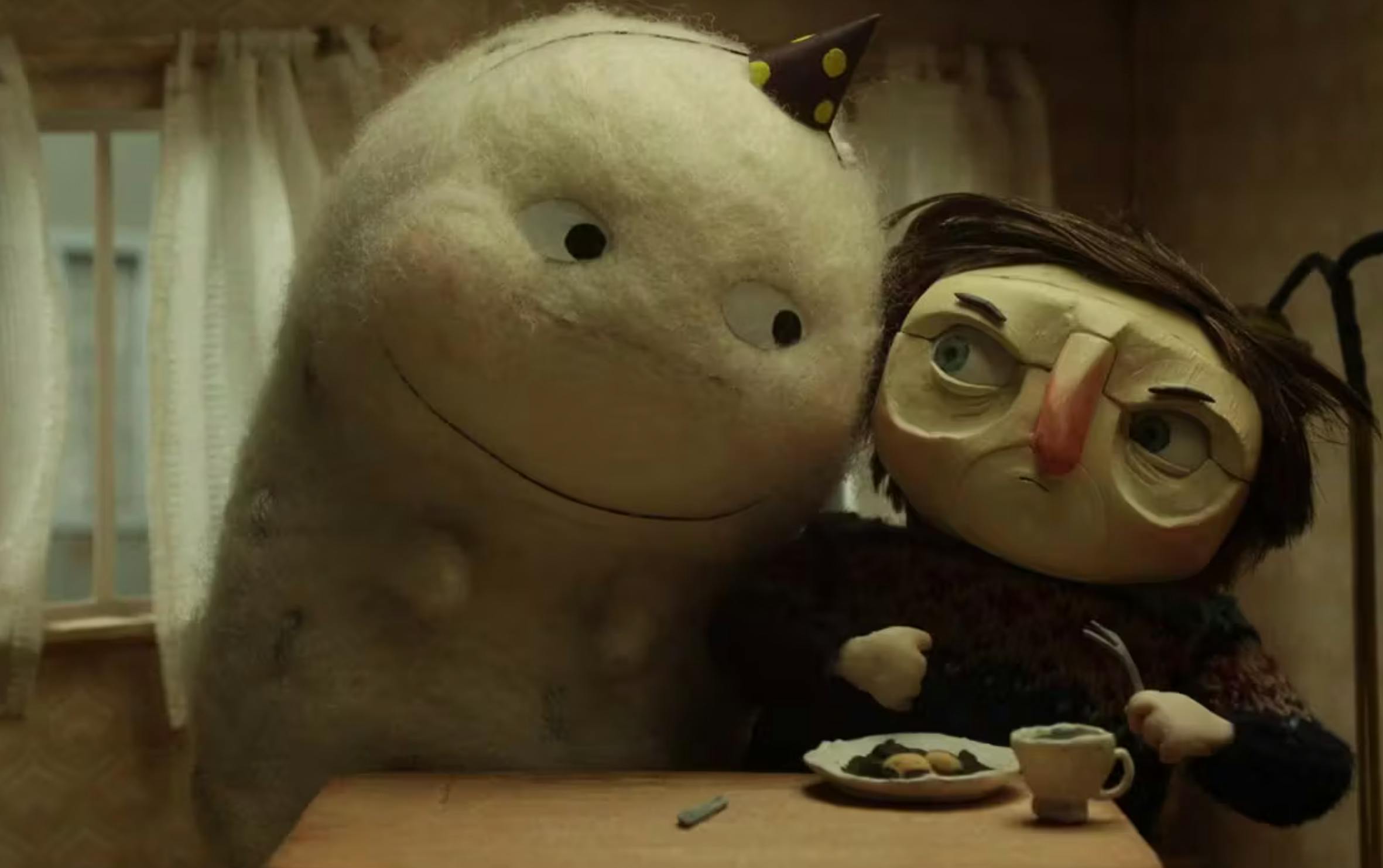 screenshot from but milk is more important (2012). A large white felted character wearing a birthday hat is looking at a human with short hair, a sweater, and is holding a fork. The human looks annoyed at the white blob character. 