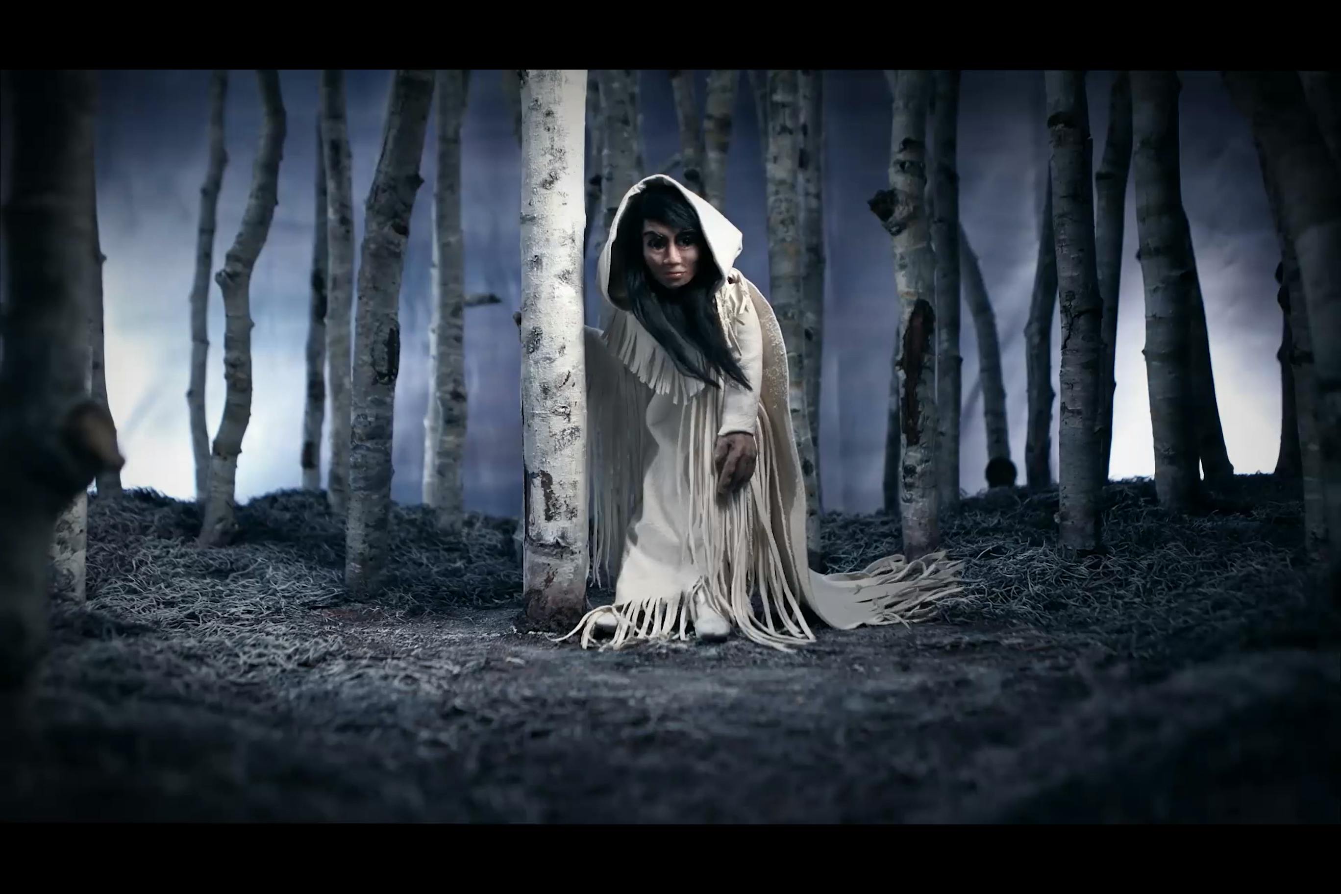 Image of  awoman in a white robe with tassels down her front stands by a birch tree and looking past the camera. The scene is in a forest