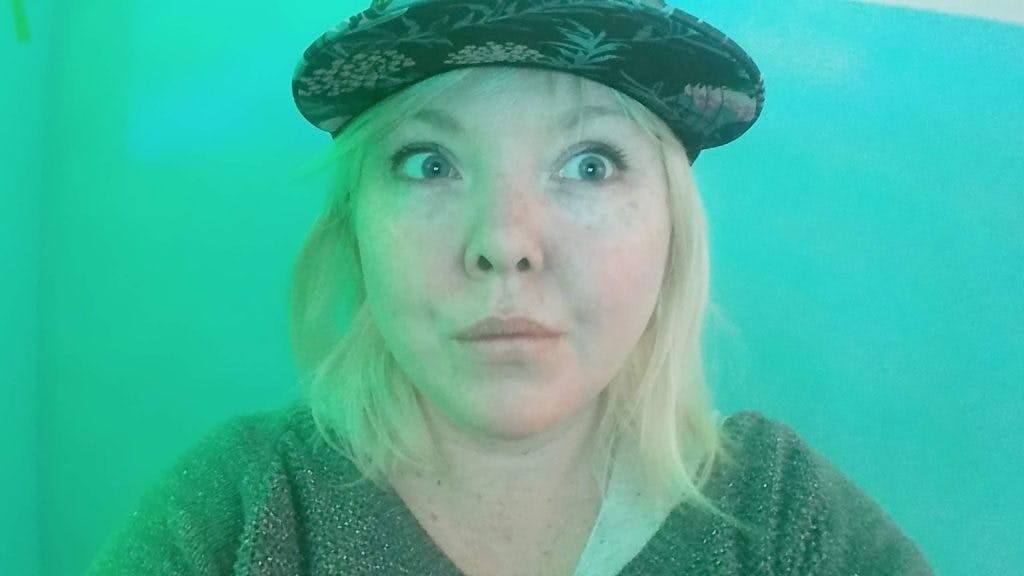 Sacha Michaud is facing the camera but looking to the right inquisitively. They're bathed in a blue-green light. They have straight, shoulder-length blond hair, blue eyes, and are wearing a pineapple-patterned baseball cap and a knit jumper