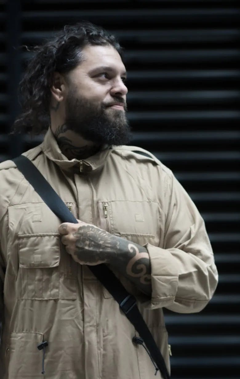 image of Tank Standing Buffalo. He is looking to the right in front of a sound-proofed wall. He is wearing a satchel, and a beige long-sleeved shirt. He has a beard and shaggy hair pinned back. He has tattoos on his hand and wrist