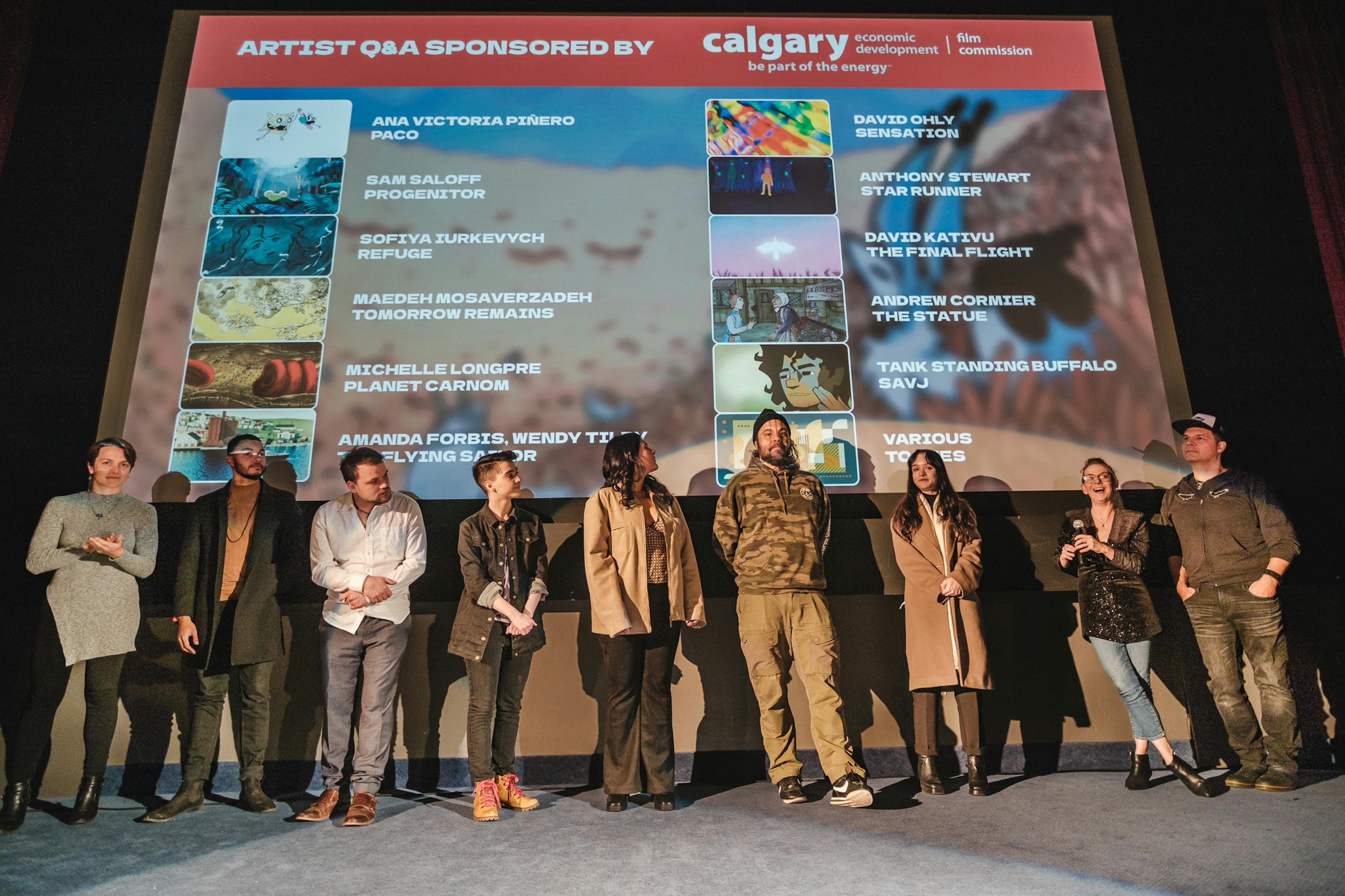 picture of 9 animators on a stage in front of a movie screen with the titles of the "Homegrown Heroes" GIRAF pack from GIRAF18. 
From left to right, Michelle Longpre, Andrew Cormier, David Kativu, Sam Saloff, Tank Standing Buffalo, Sofiya Iurkevych, Troy Kokol