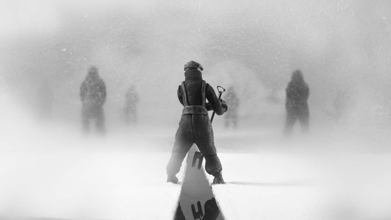 A figure in heavy winter clothing straddles a gap in the ice, as similar figures stand in the background obscured by falling snow.