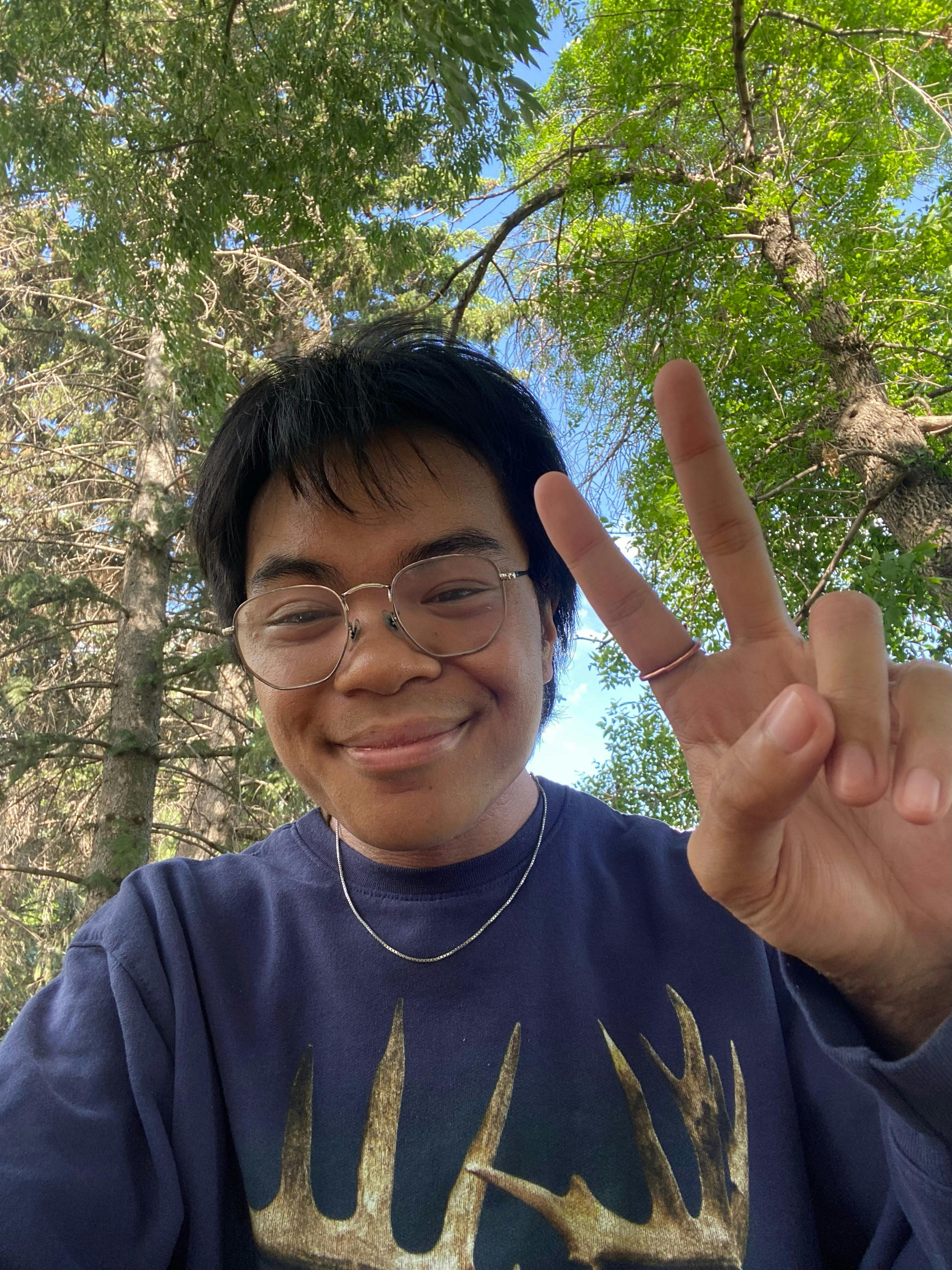 headshot of Daniel Volante. Daniel has short black hair, wears square-rimed glasses and is wearing a thin silver necklace. He is wearing a blue pull-over sweater with antlers on it. Daniel is smiling and holding up his left hand with the "peace" sign. His index finger has a thin gold ring on it