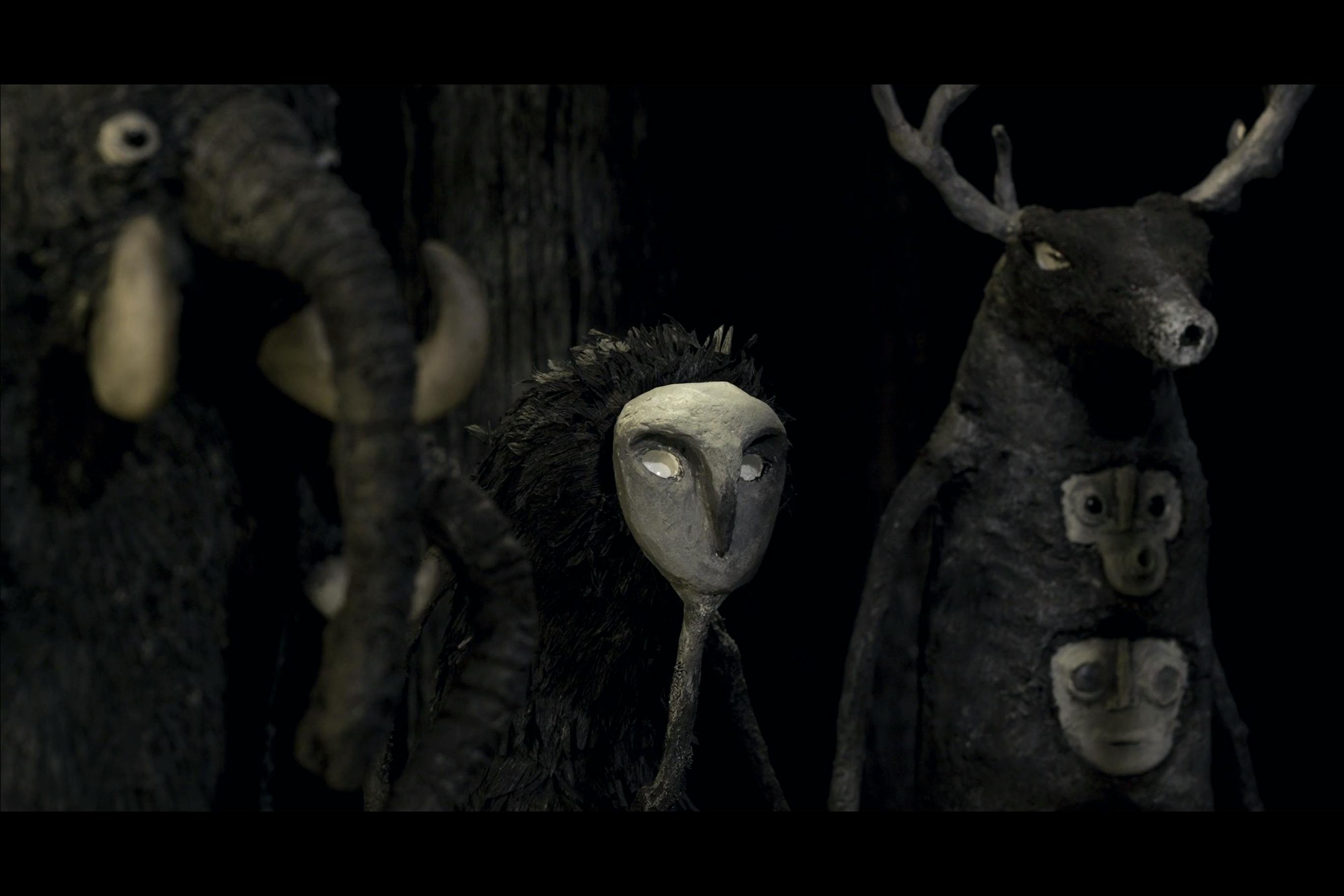Three characters are looking off-screen to the right. The leftmost character is a character with an elephant face, and elephant faces for arms. The middle scaracter looks lik ea furry being wearin gan owl mask. The third character has antlers, and three faces stacked on top of each other like a Totem pole