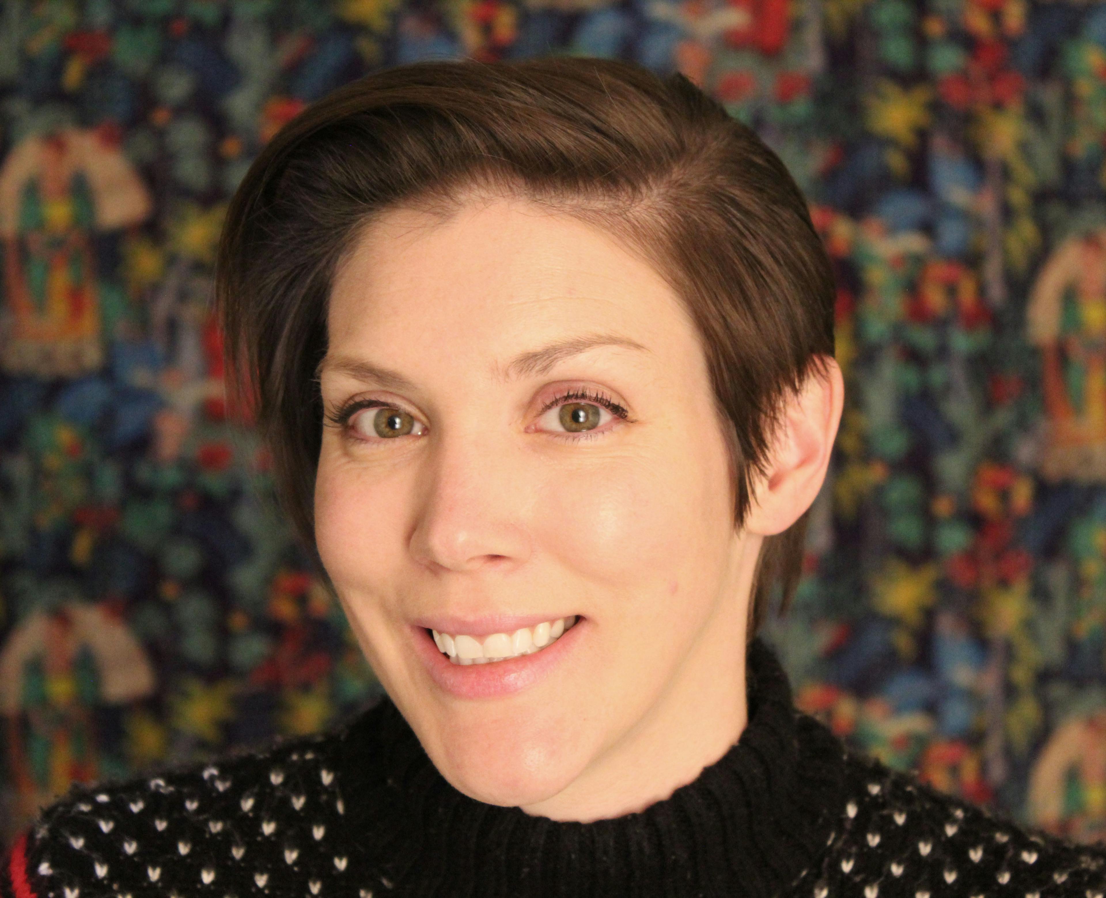 Headshot of Val Duncan. Val has short brown hair with long bangs brushed to the side. Her eyes are hazel, and she is smiling. She is wearing a knitted jumper that is black with white speckles. 