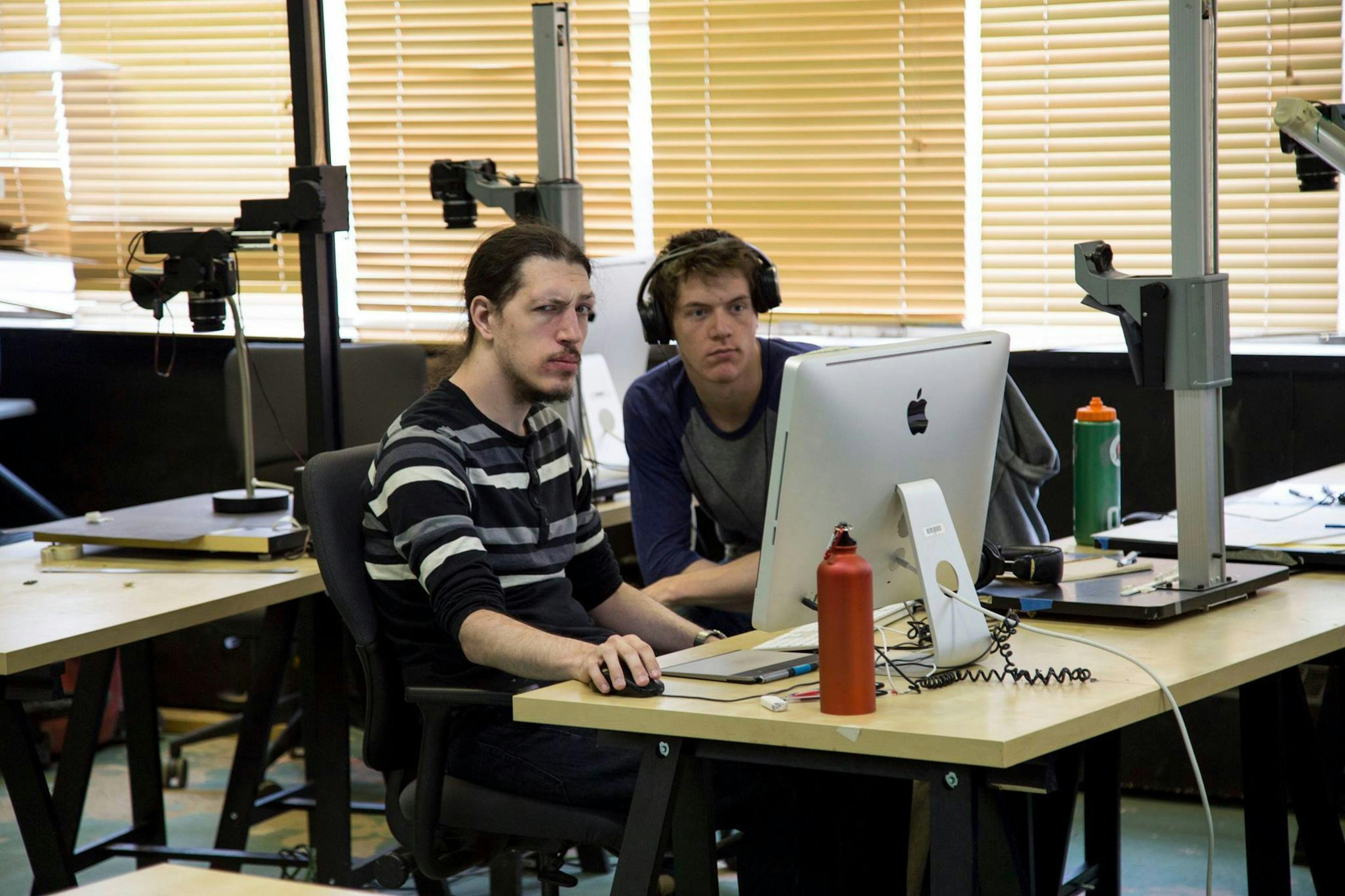 Will Walton and another animator sitting at a desk with an iMac and an animation stand. Will has long hair that is tied into a pony tail at the base of his neck. He has a moustache and beard, and is wearing a striped black and white shirt. He is looking at the camera with a raised eyebrow. Next to him the other animator is looking studiously at the computer while wearing headphones 