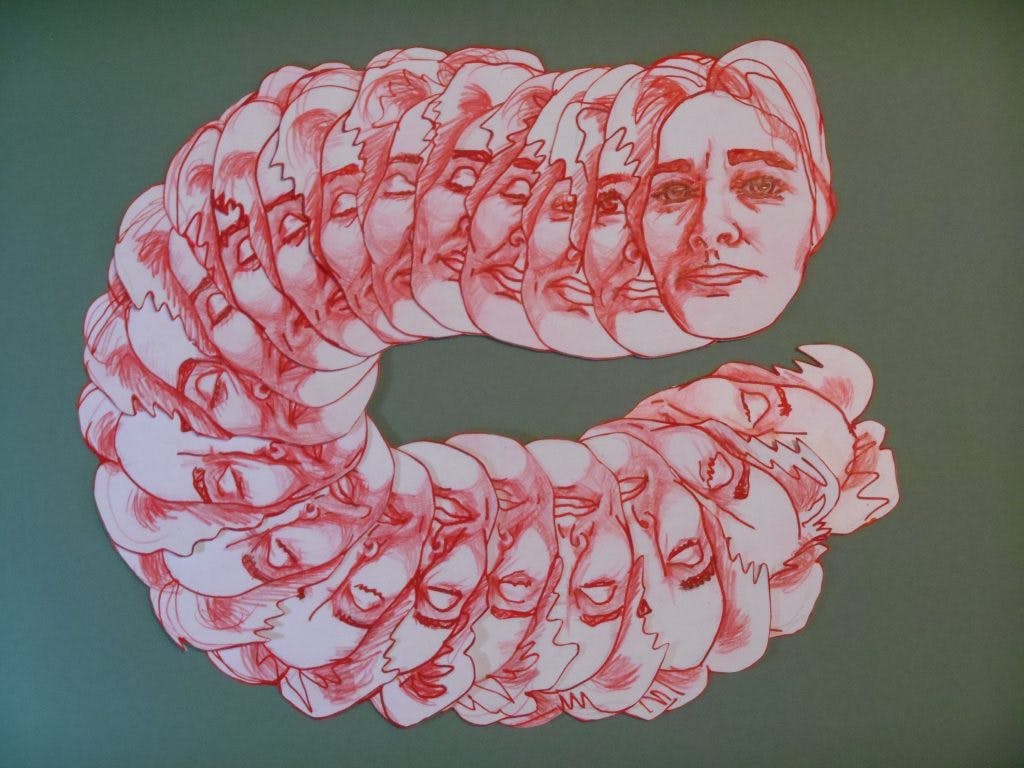 a picture of multiple paper-cut-outs of a face blinking. The paper cut outs are overlapping each other and going in a "C" shape. They are drawn in red pencil and are laying on a green tablecloth