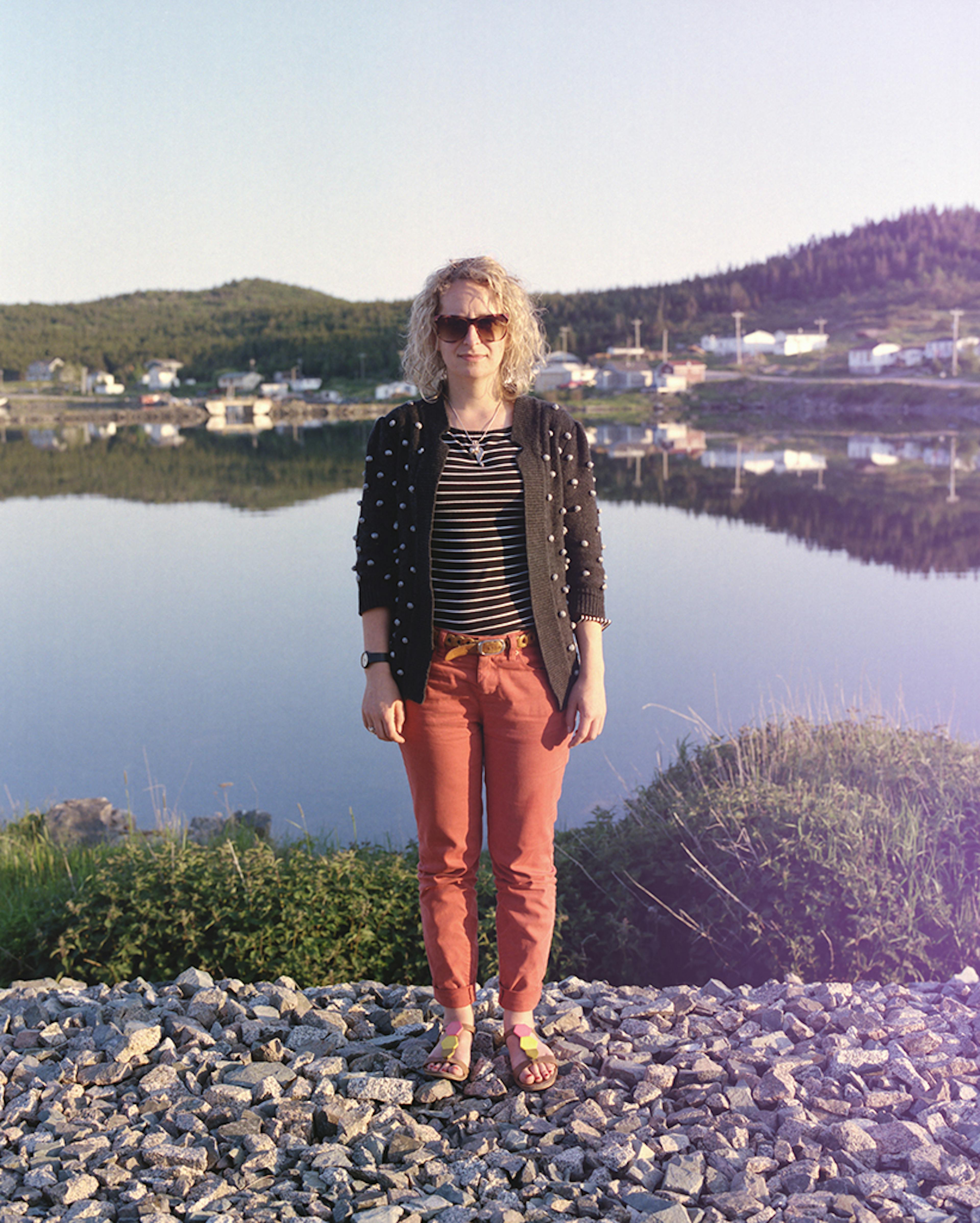 image of Yvonne Mullock in front of a backdrop of ocean and houses. She has curly shoulder-length hair, and is wearing sunglasses, a striped top with a black-and-white polka-dotted cami, and red pants. 