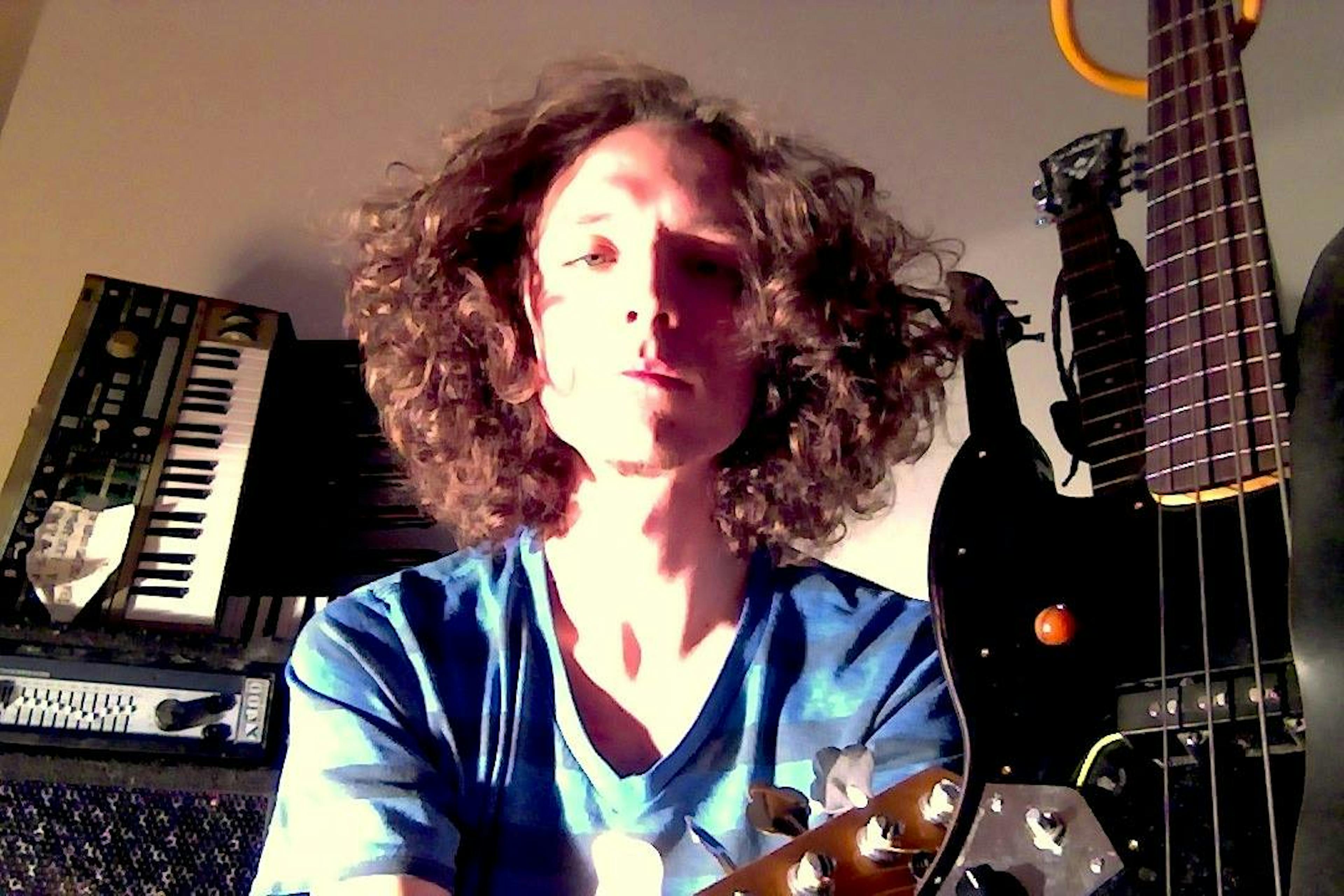 Headshot of Eric Smith - a person looking down towards the camera, they have curly hair that reaches the nape of their neck. They're wearin ga blue striped t shirt and are surrounded with musical instruments including an amp, an electric guitar, and an electric keyboard