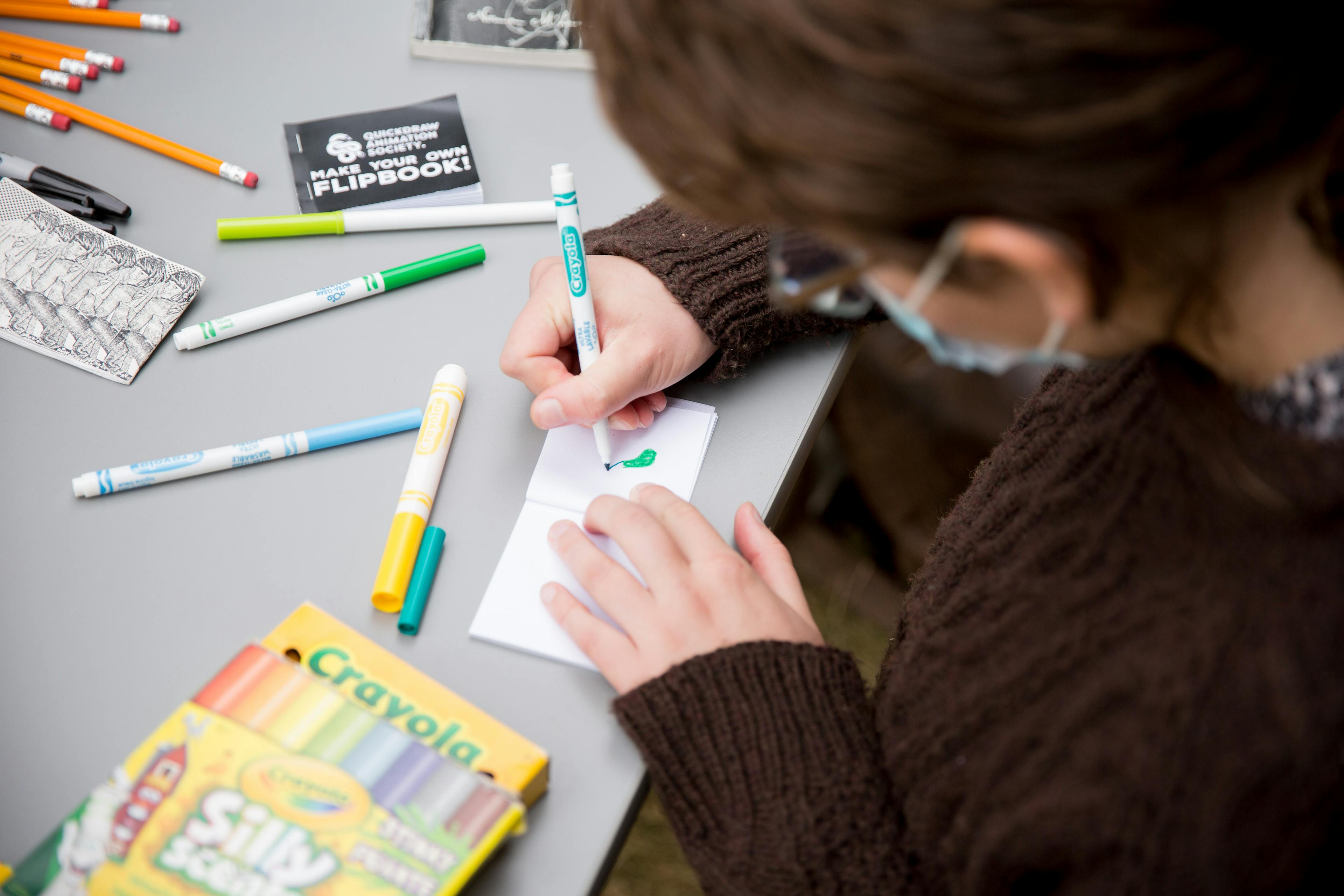 Photograph of a person from behind their left shoulder. 

Person is wearing glasses, a brown cabled-knit sweater and a face mask. 

They are sitting at a table that's covered in colored markers and a book that says "quickdraw animation society make your own flipbook!"

They are drawing something in green on a flipbook page