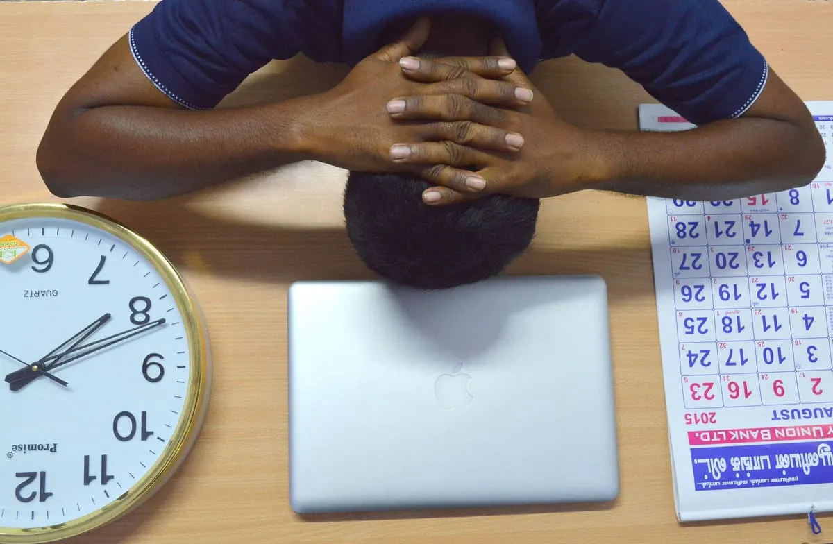 A man resting his head on his hands at a desk beside a clock